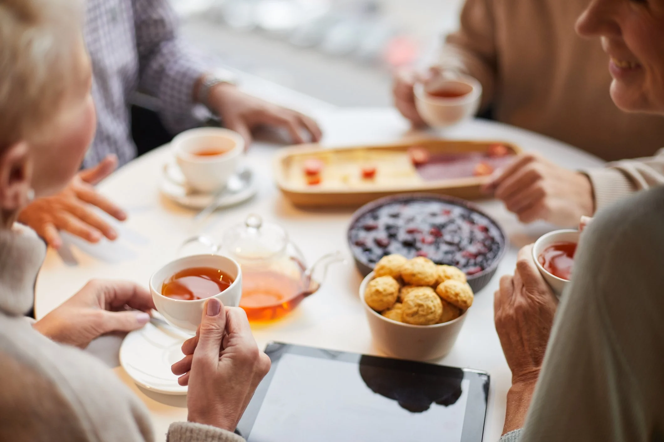a group of people drinking tea at a table with trays of pastries and a glass of teapot
