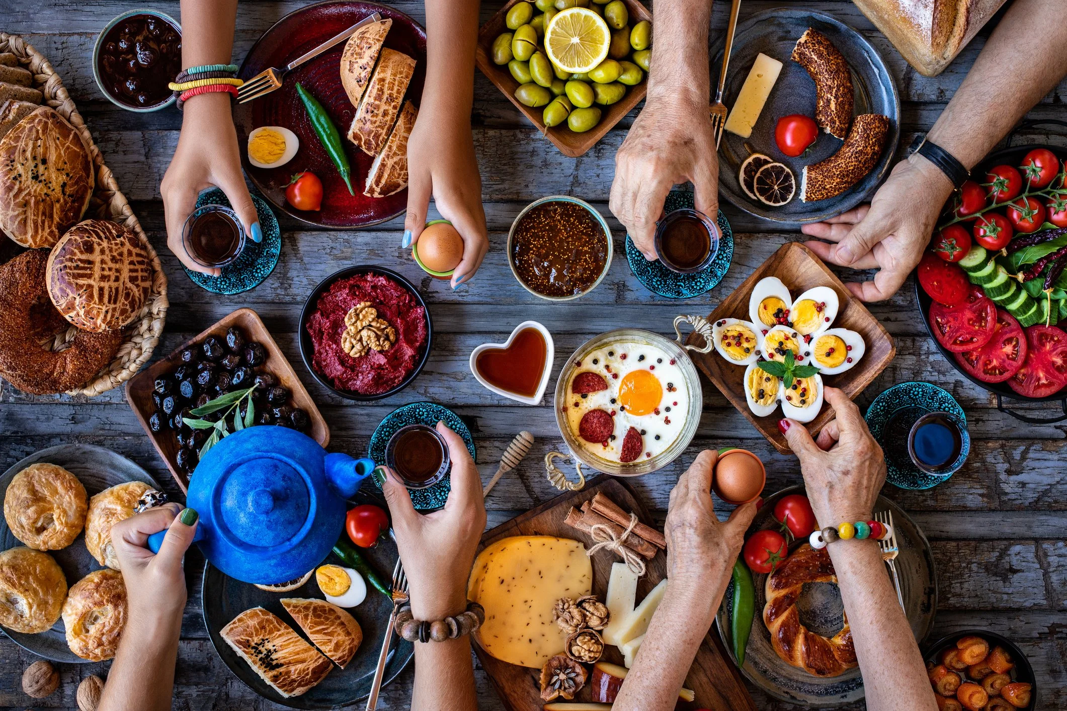 Overhead view of friends sharing tea and a colorful brunch spread with pastries, eggs, cheese, olives, and vegetables.