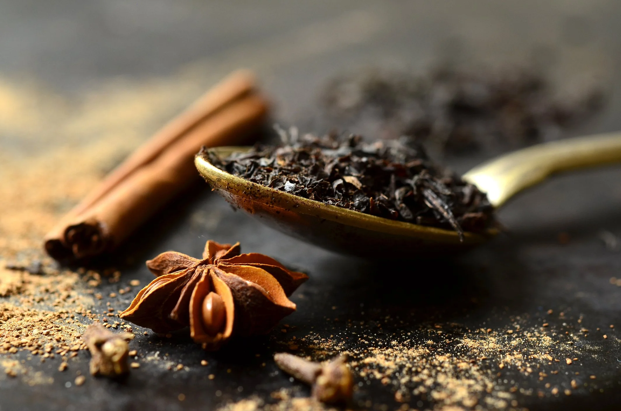 A spoon holding black tea leaves on a table next to cinnamon and star anise