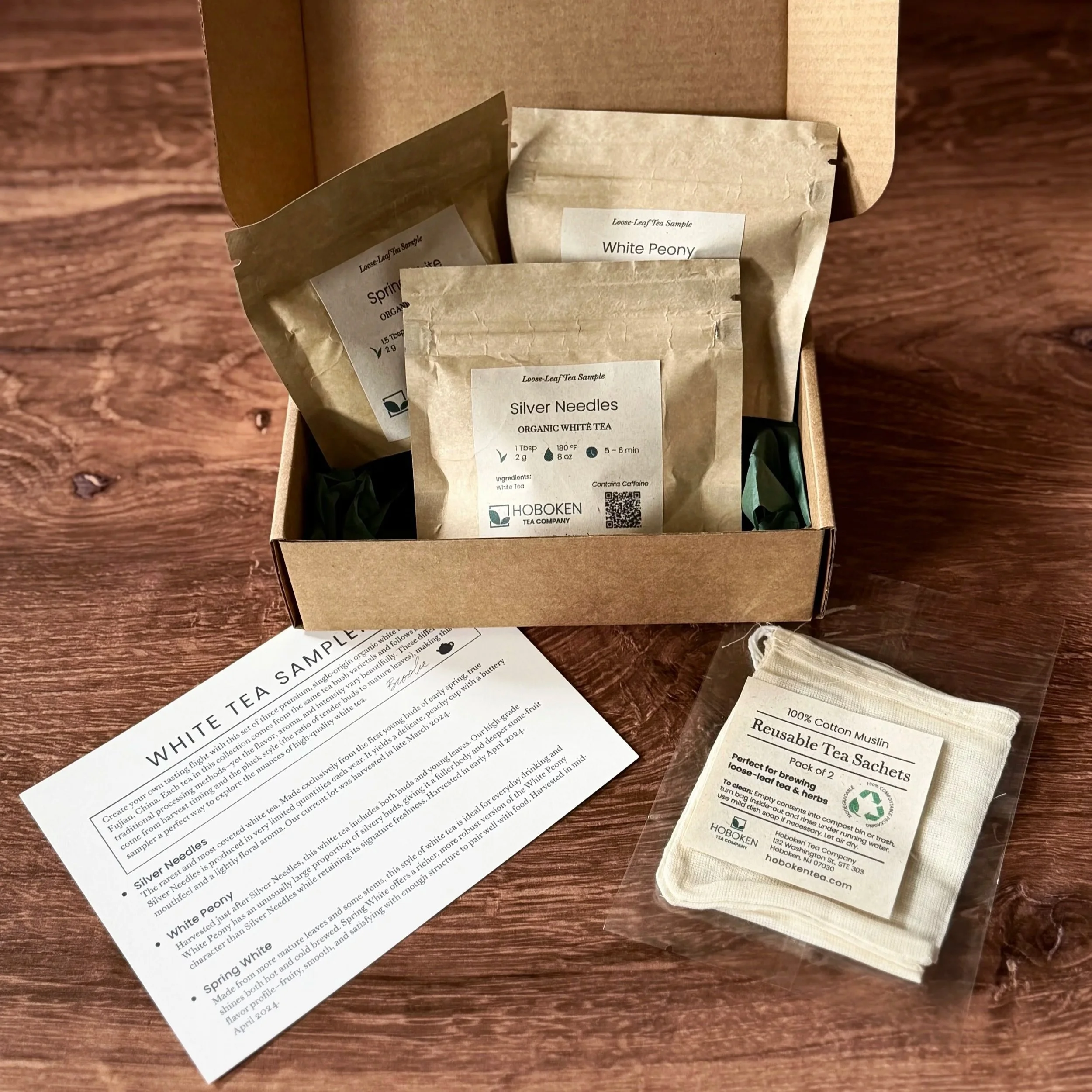 An open box containing 3 small packets of loose leaf tea on a table next to cotton tea bags and an info card