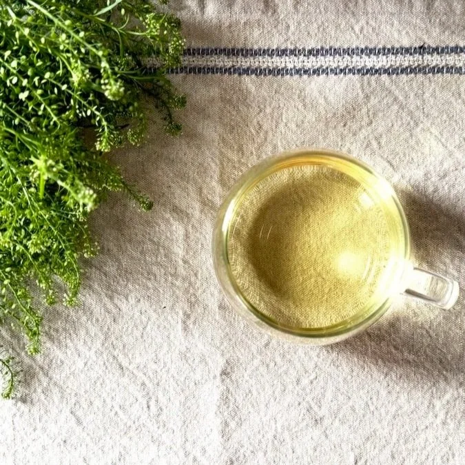 a glass cup of green tea on a linen table runner next to a sprig of green leaves