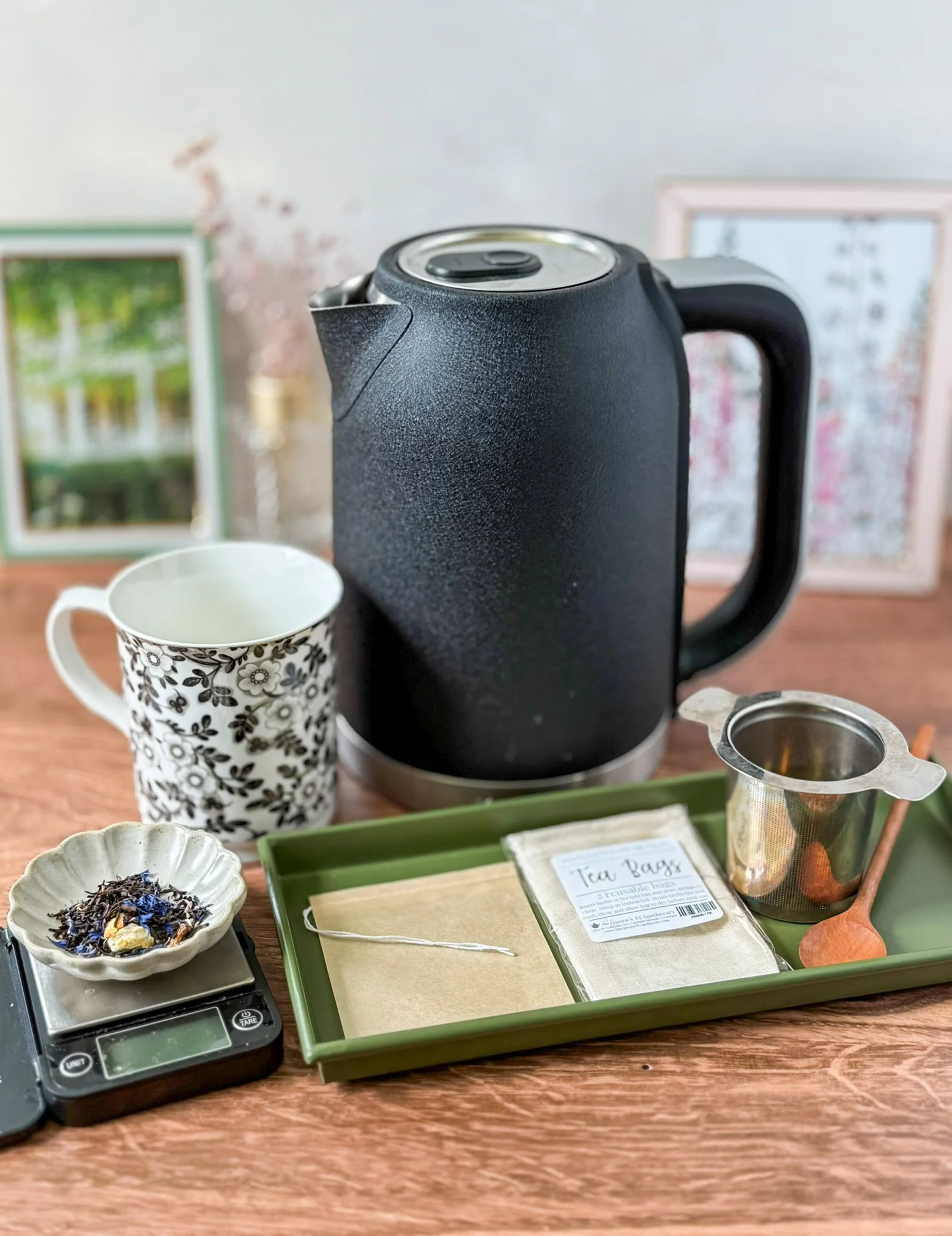 an electric kettle on a table next to a mug, a dish of dry tea leaves on a scale, and a tray with tea brewing implements