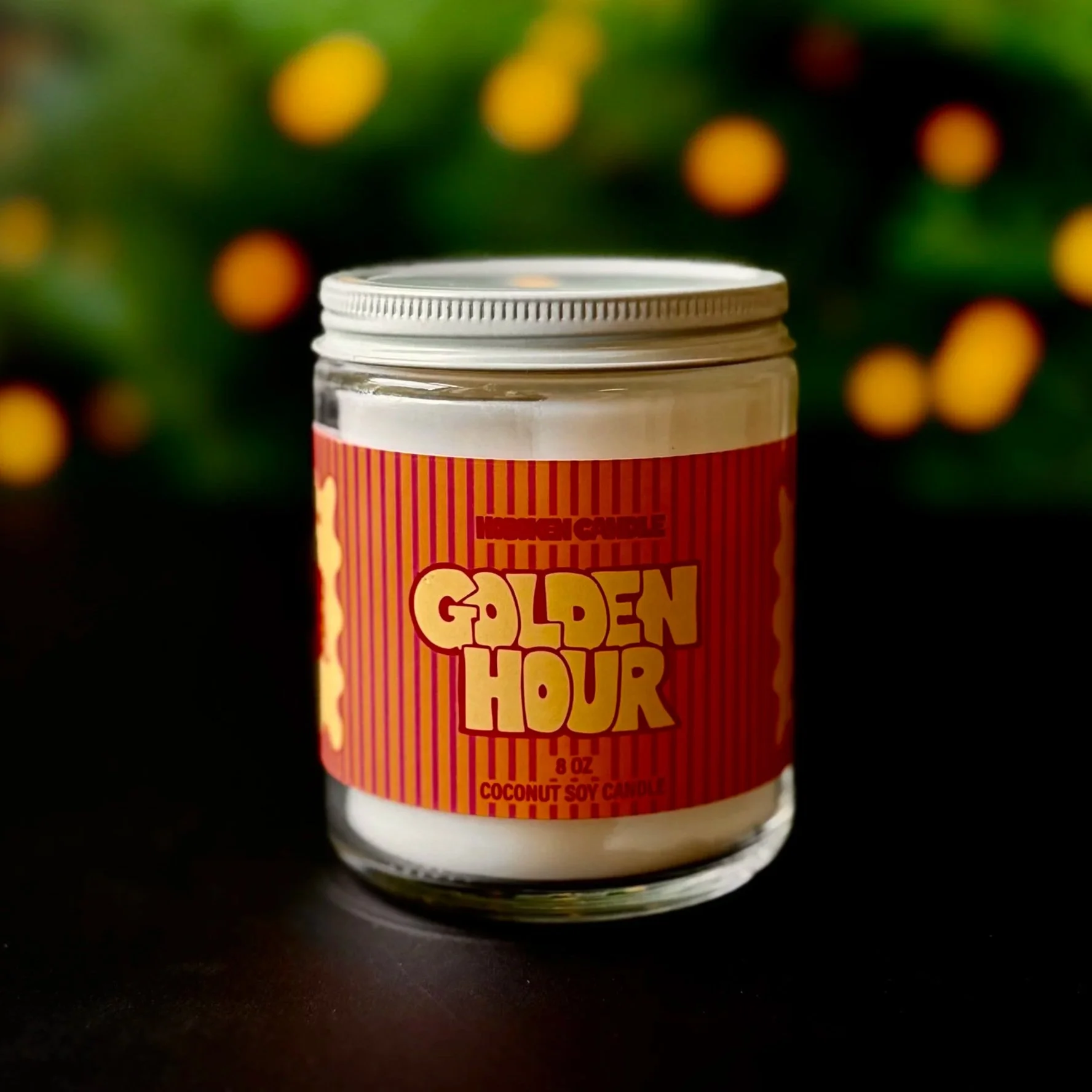 A glass jar labeled Golden Hour with a Hoboken Candle label in red and yellow on a black surface with lights and greenery in the background