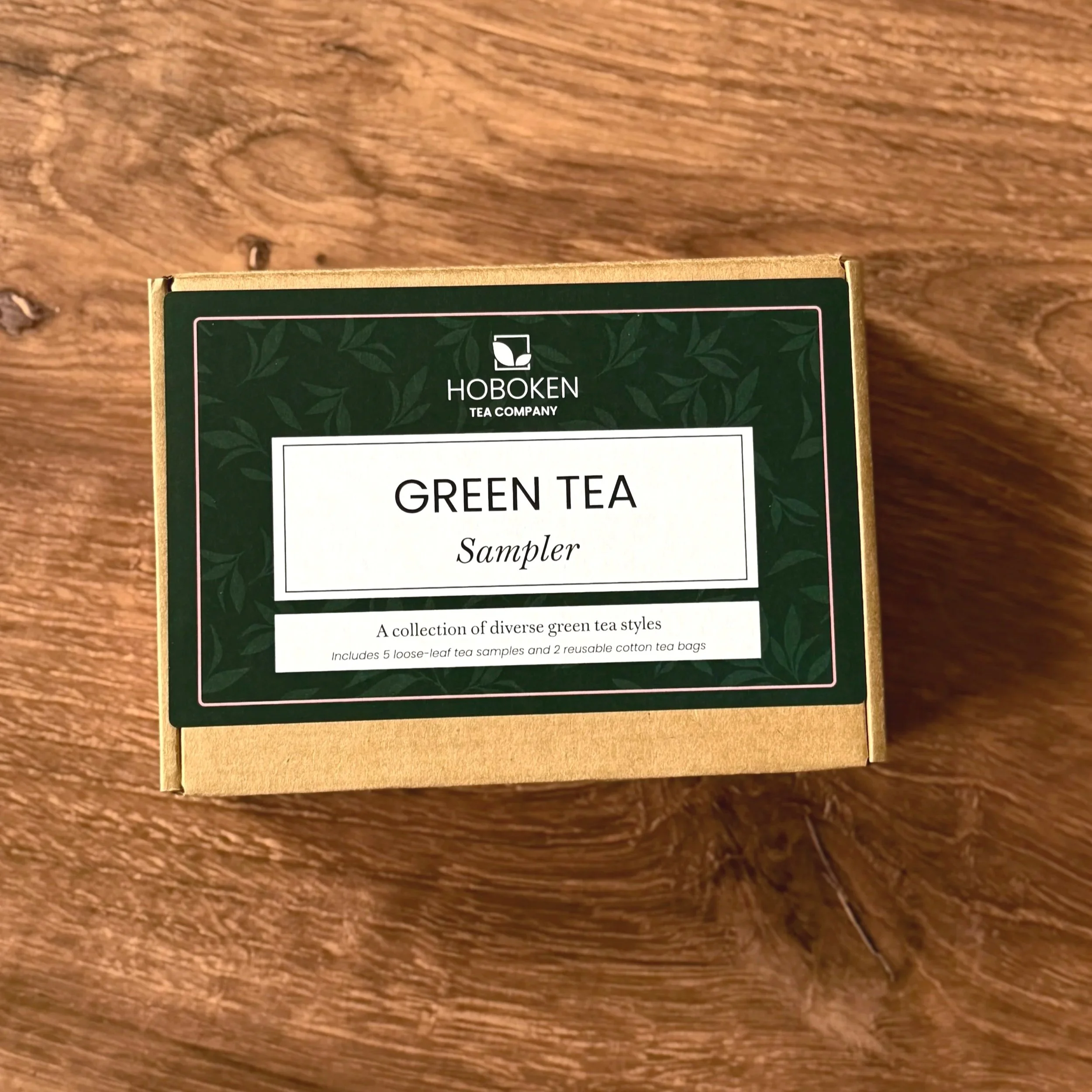 A box with a green Hoboken Tea Company label reading "Green Tea Sampler"  on a wood table