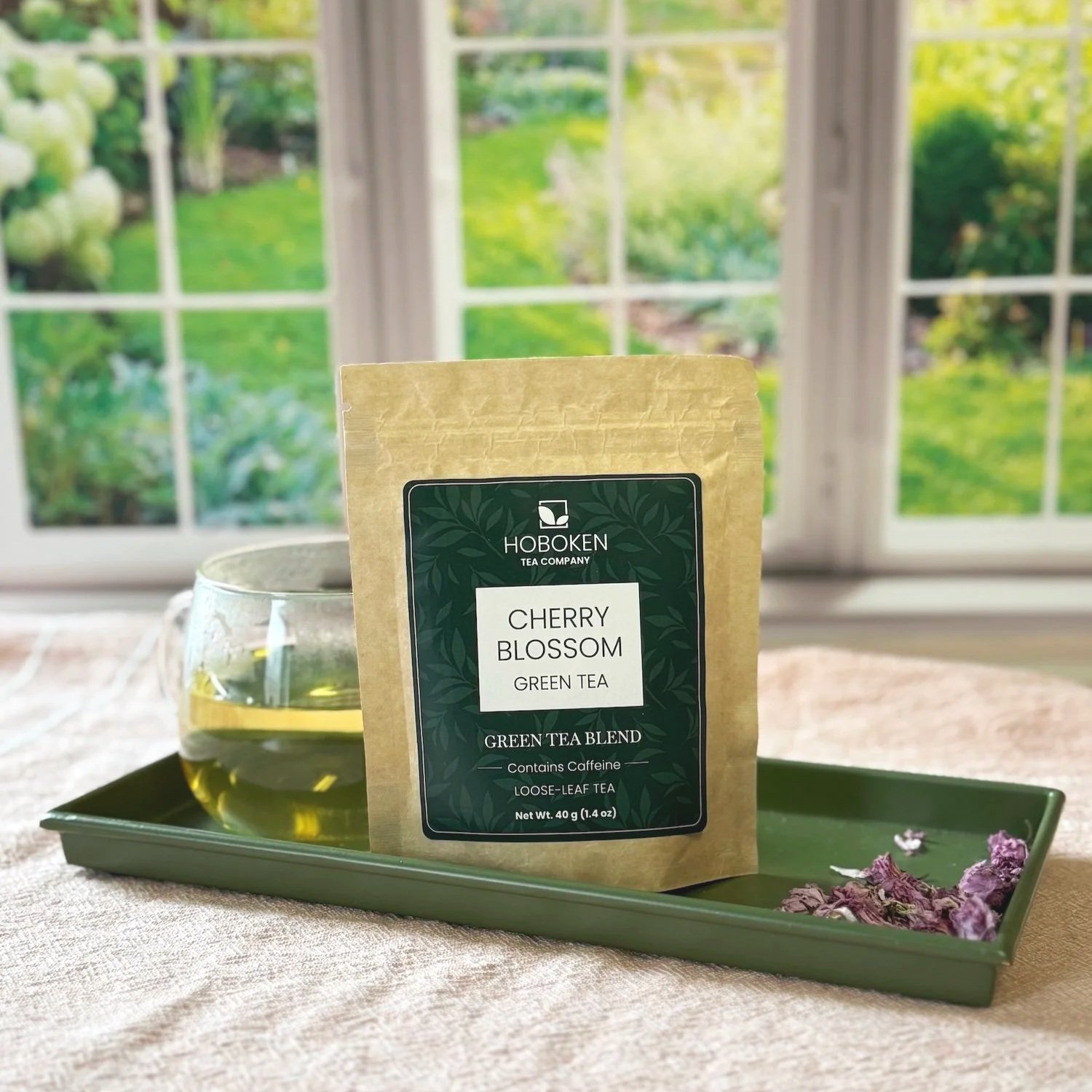 kraft paper pouch of Cherry Blossom Green Tea on a tray next to a cup of green tea in front of a window with a garden outside