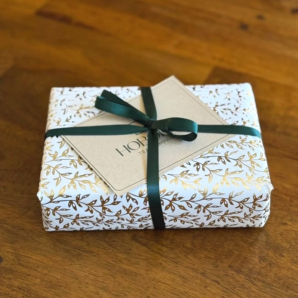 a package wrapped in gold leaf pattern wrapping paper with a green ribbon and kraft paper note card on a wood table