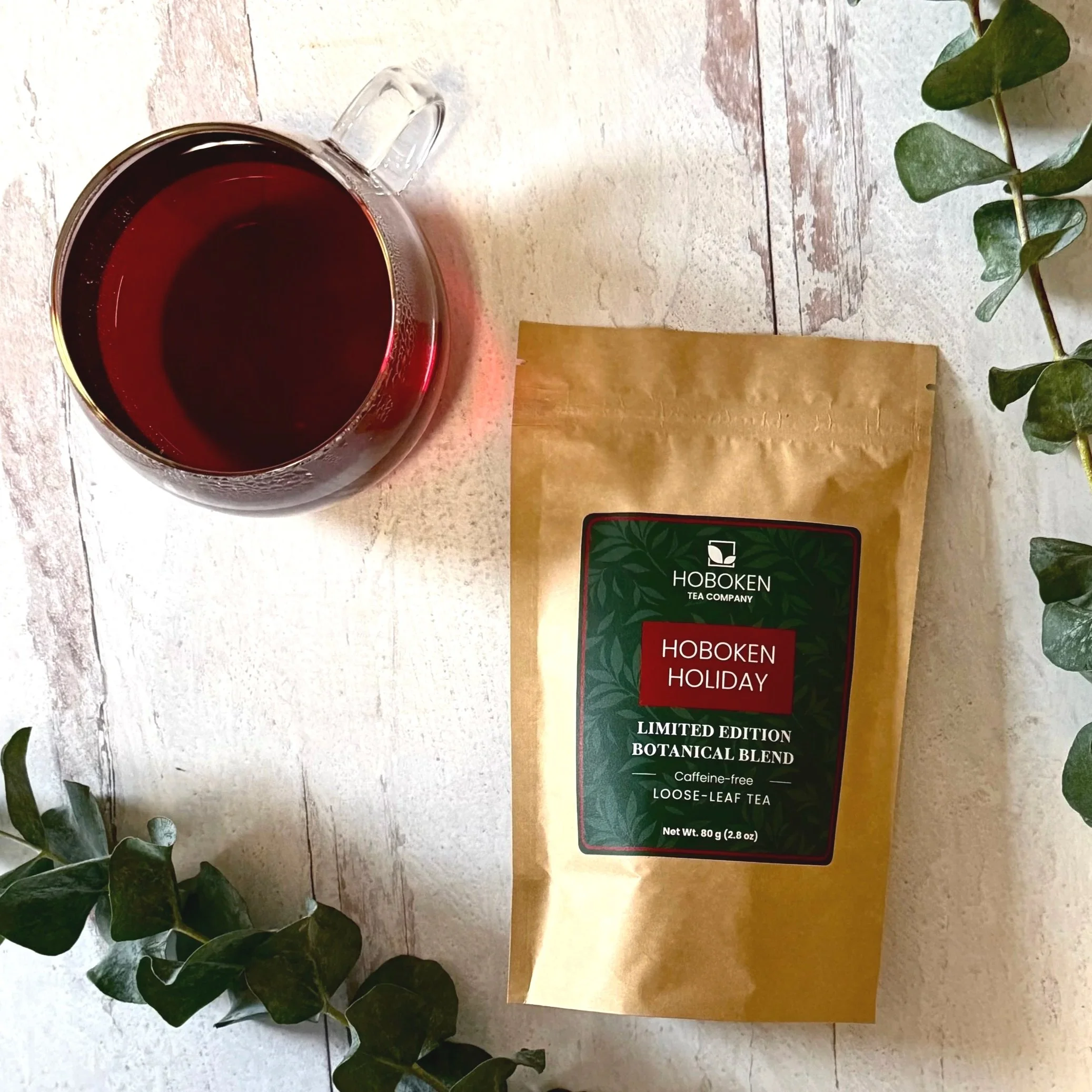 a package of Hoboken Holiday caffeine-free loose leaf tea from Hoboken Tea Company on a white wood surface next to a cup of dark red tea and some green leaves
