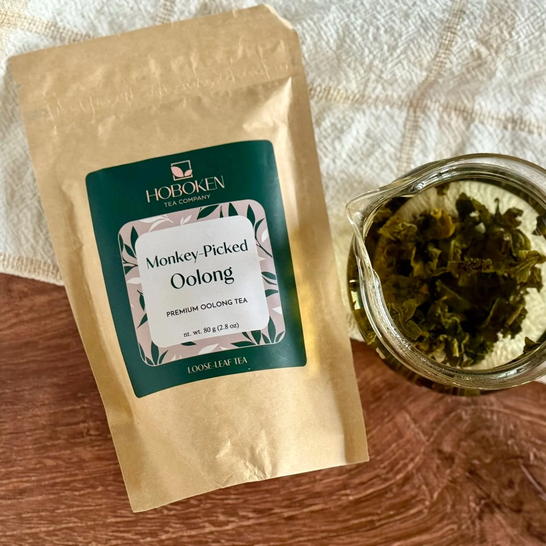 Package of Monkey-Picked Oolong with Hoboken Tea Company label next to a glass teapot filled with water and green oolong tea leaves