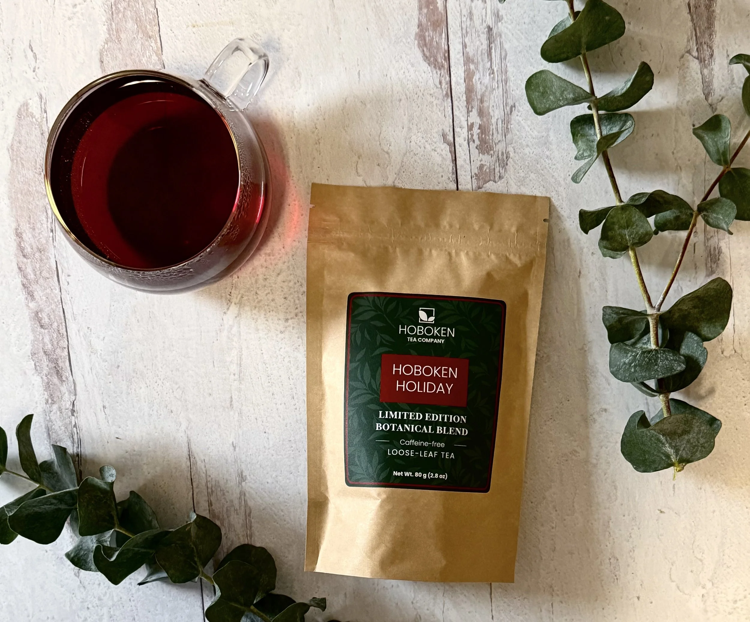 A package of Hoboken Holiday loose leaf tea from Hoboken Tea Company on a light wood table next to a cup of red herbal tea