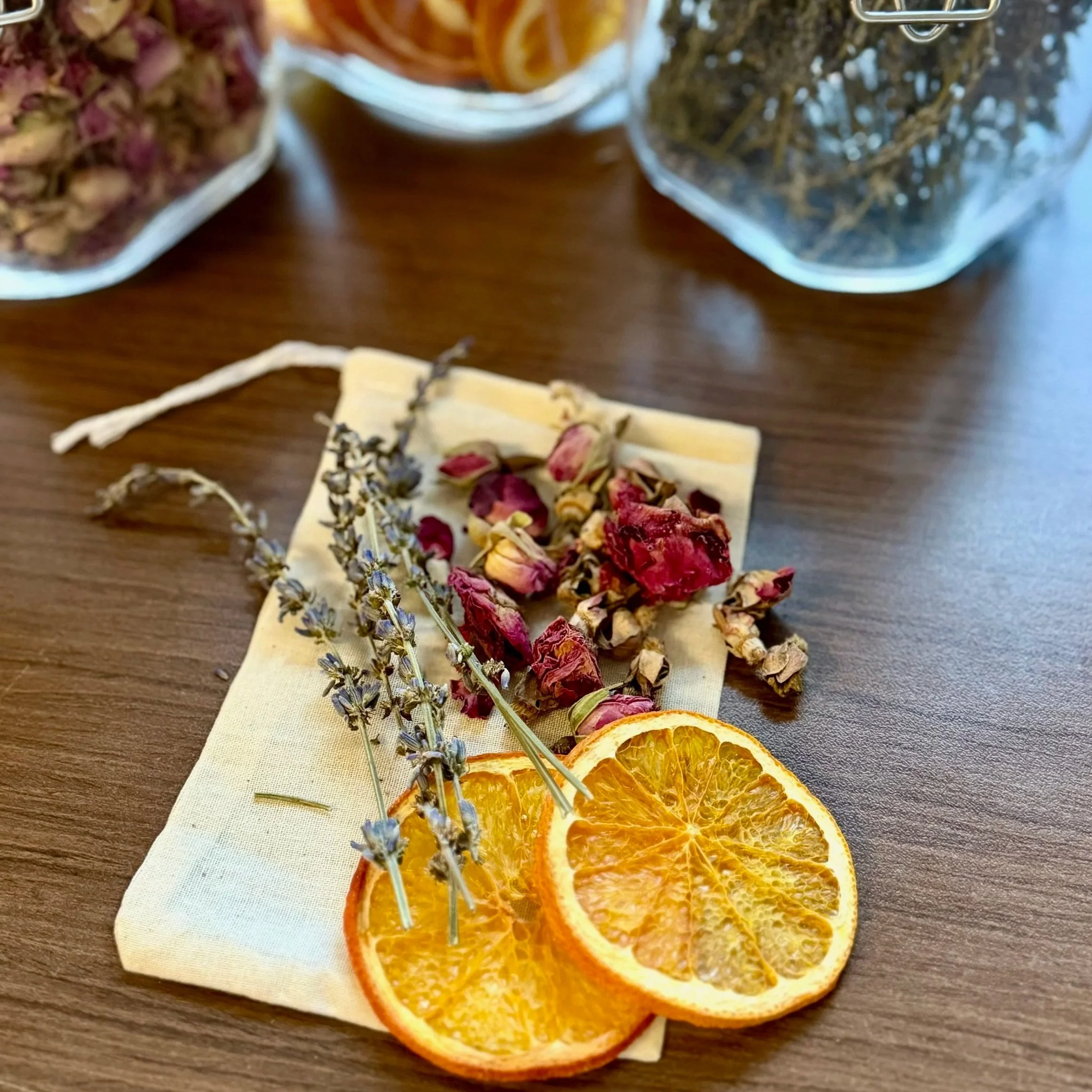 dried rosebuds, lavender, and orange slices on a muslin bag on a table next to jars of dried herbs