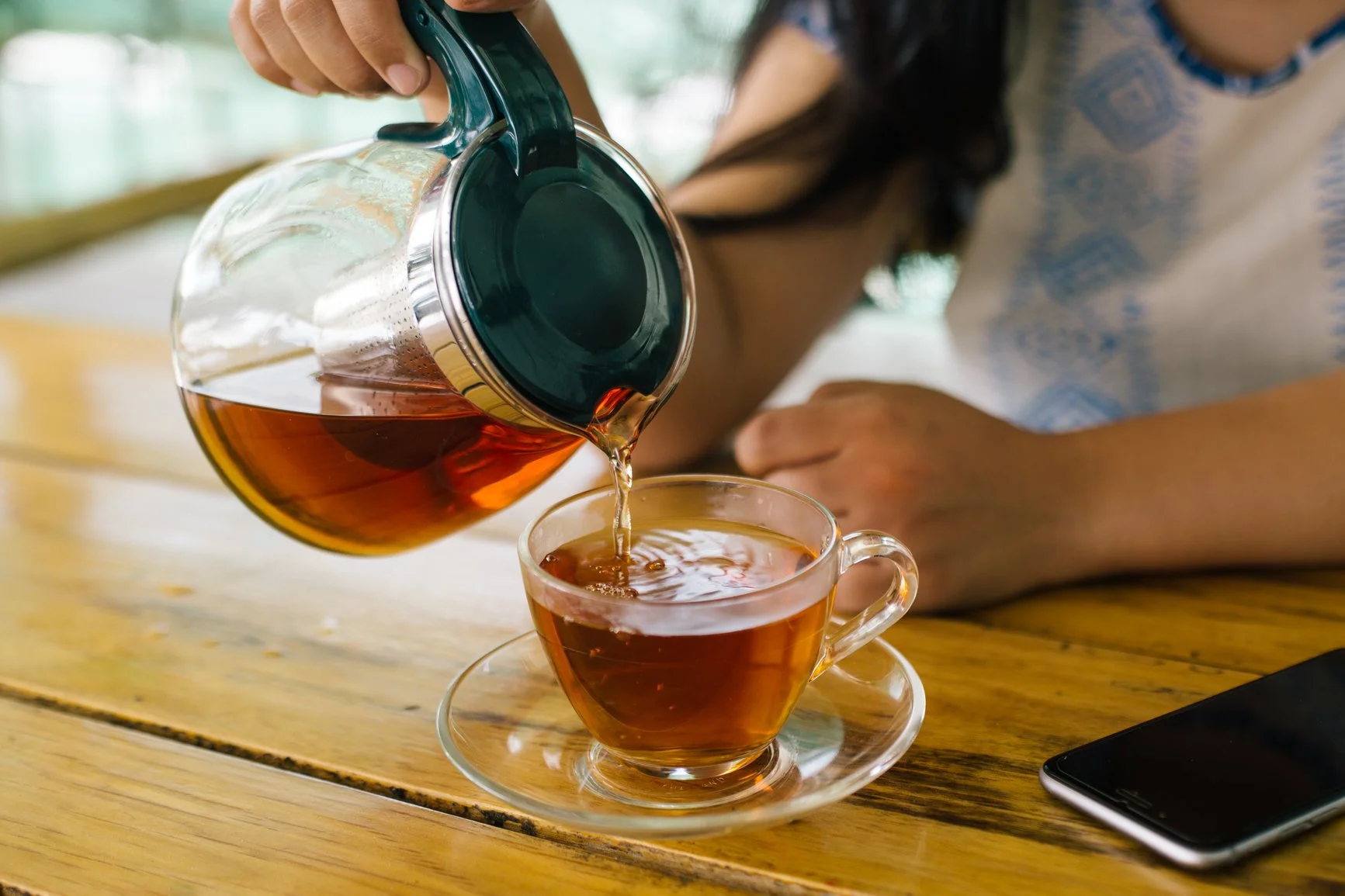a woman pouring tea from a glass teapot into a cup on a wooden table next to a smartphone