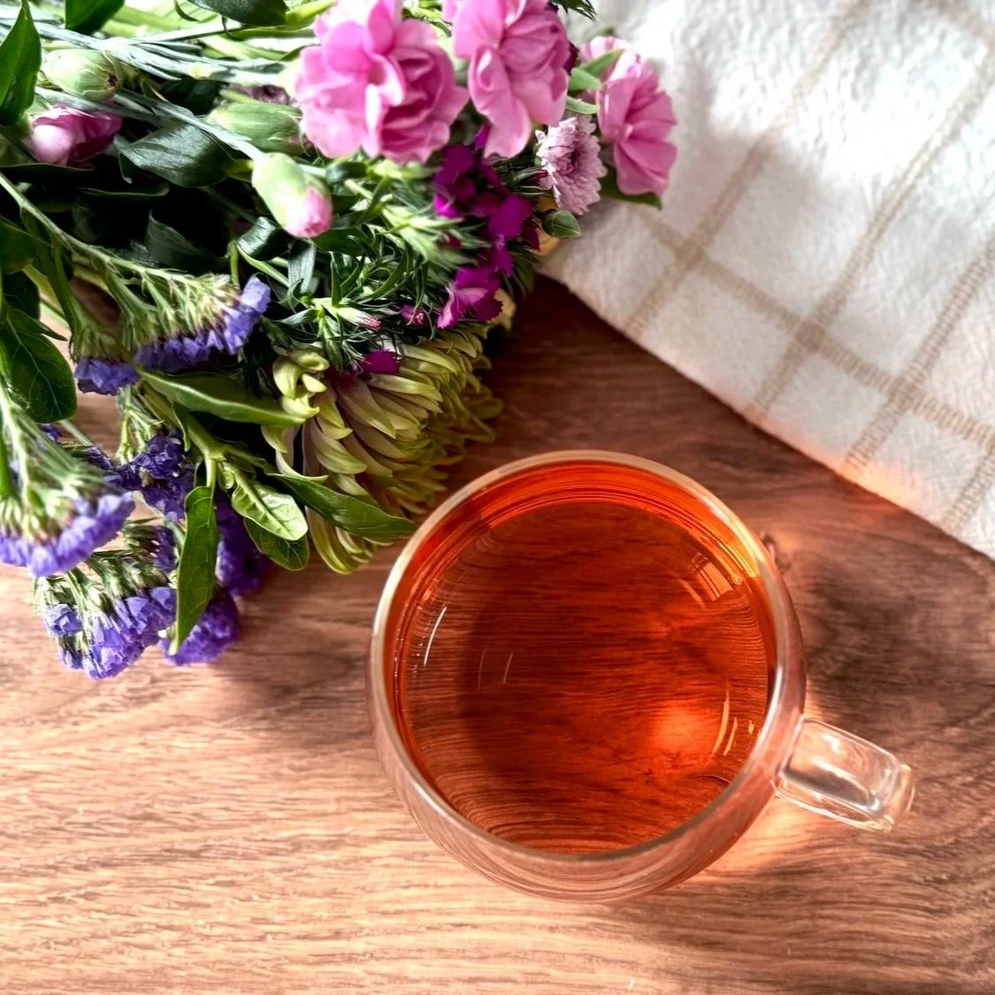 a glass cup of herbal tea on a wood table next to a bouquet of pink and purple flowers