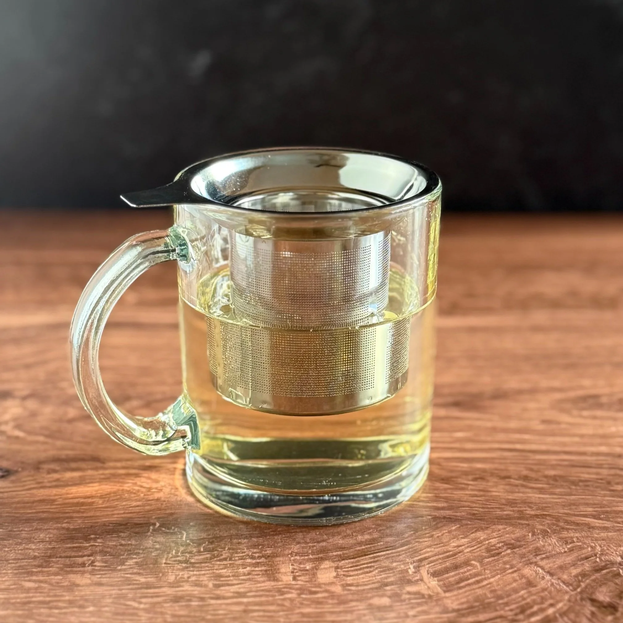 a glass mug of green tea with a metal infuser basket set inside, on a wooden surface