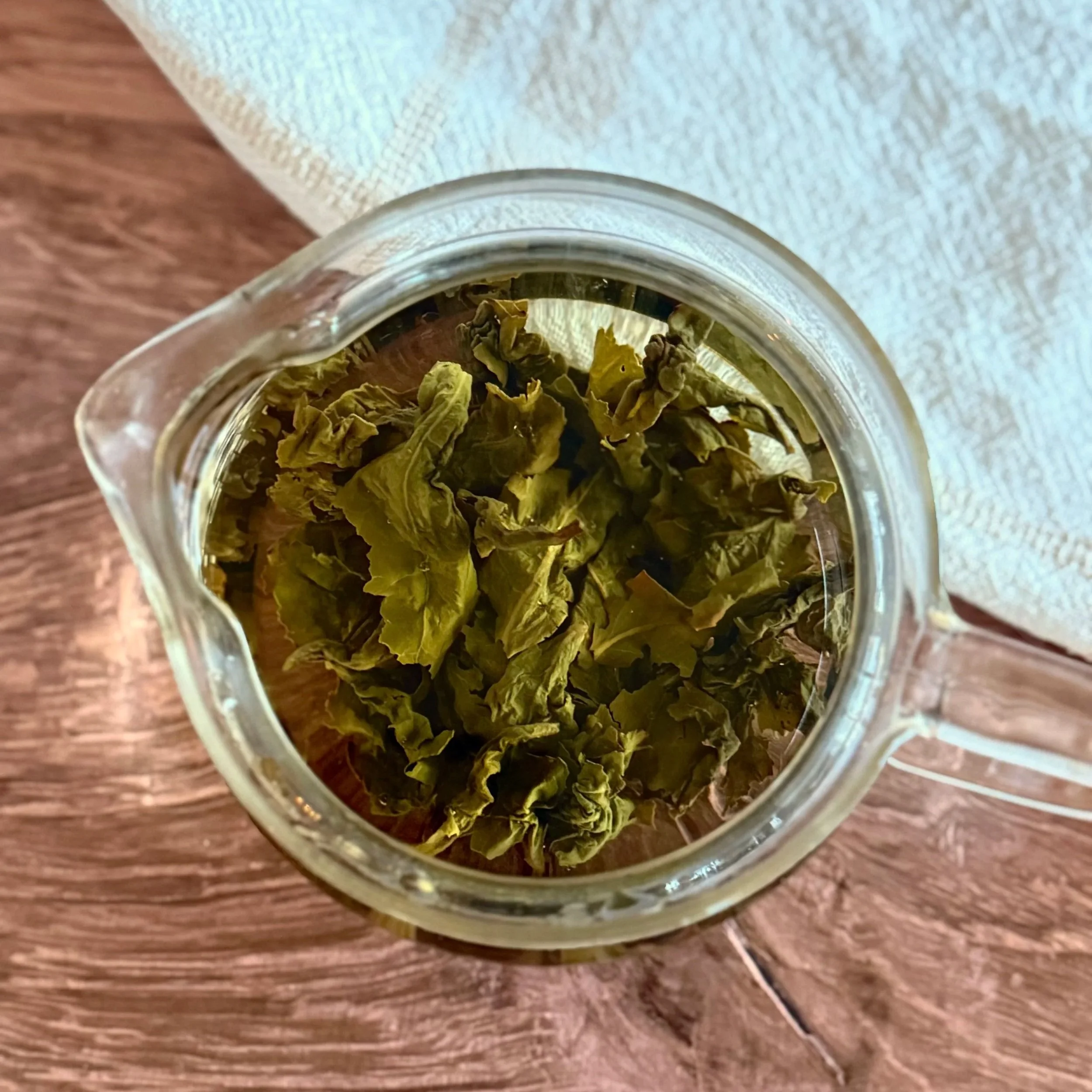 A glass teapot filled with water and large green oolong tea leaves on a wood background