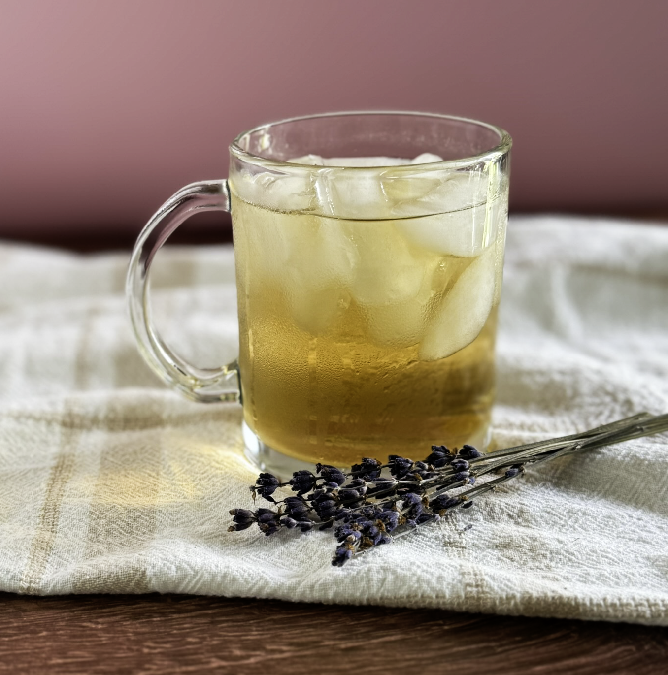 a glass mug of lavender chamomile iced tea next to a sprig of dried lavender on a linen cloth