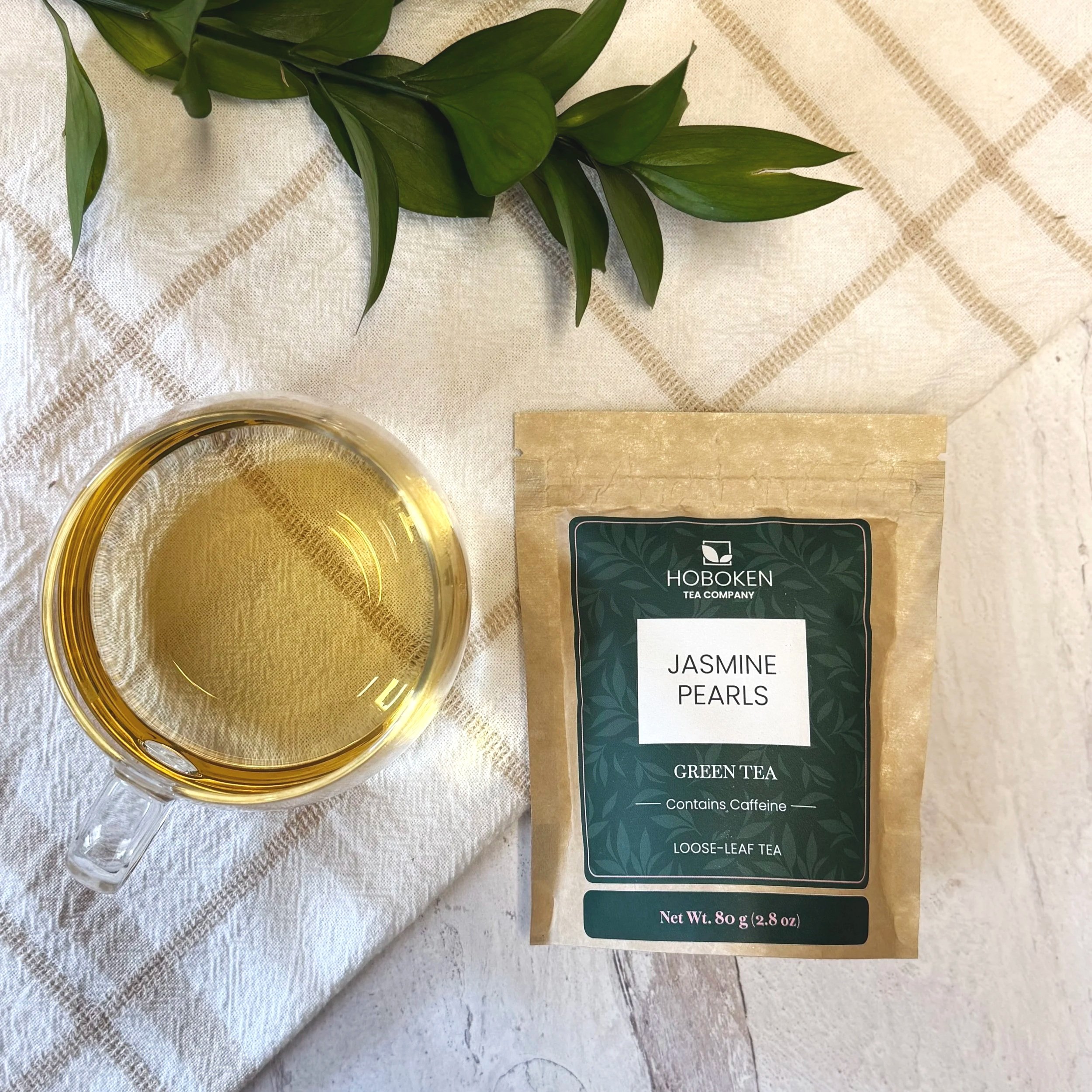 a package of loose leaf Jasmine Pearls green tea from Hoboken Tea Company next to a glass of green tea and some green leaves