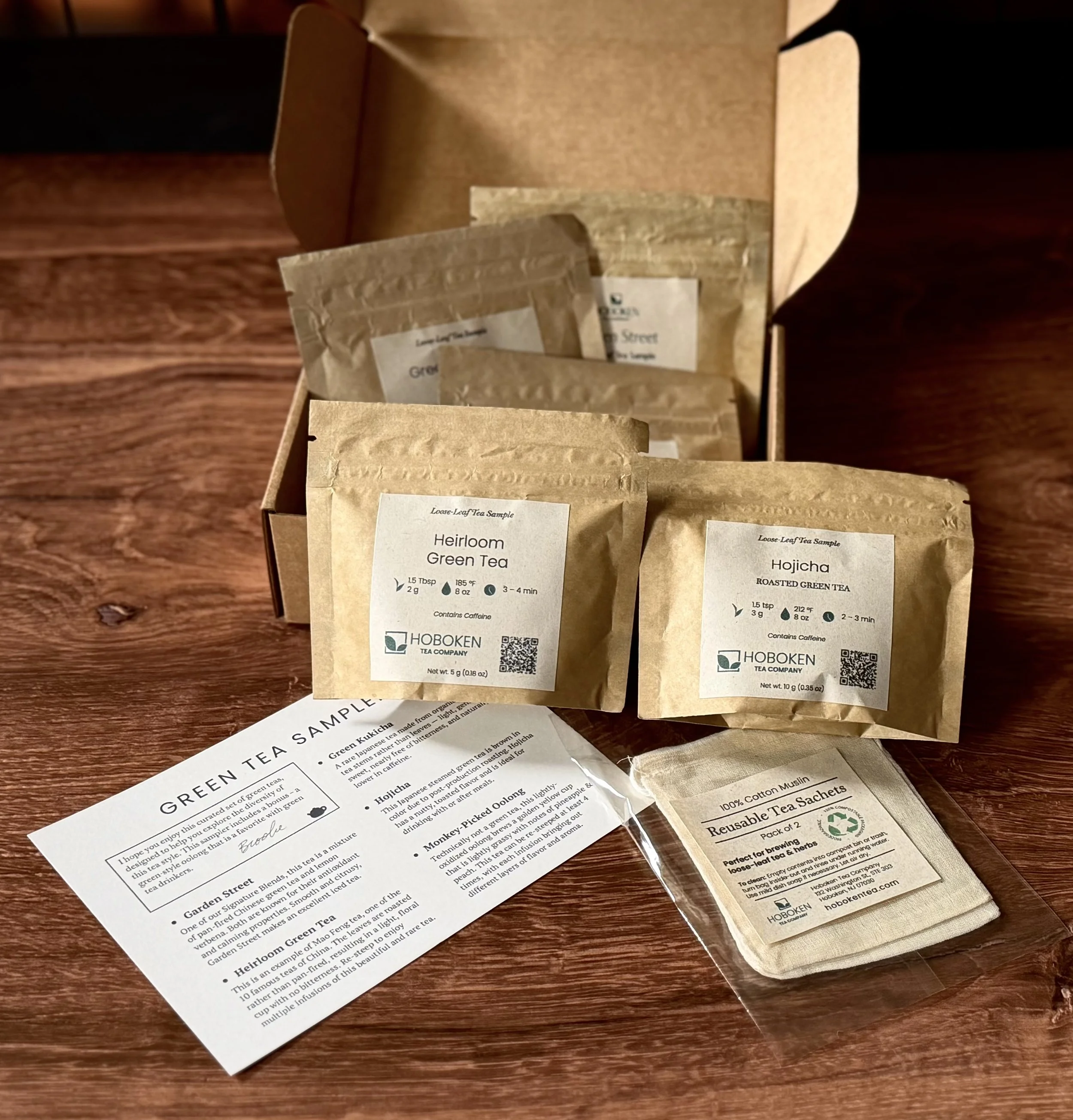 An open box containing several small packets of loose leaf tea on a table next to a pack of reusable cotton tea bags and an info card