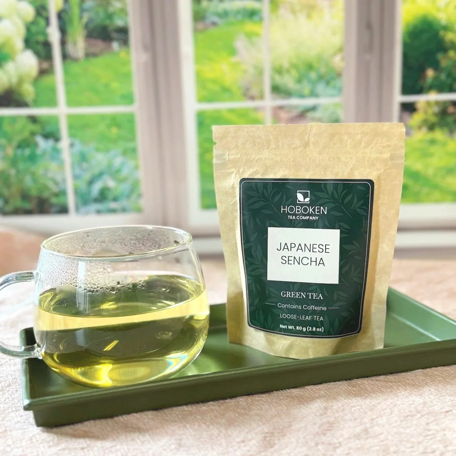 a paper packet of loose leaf sencha green tea on a tray next to a cup of green tea in front of a window