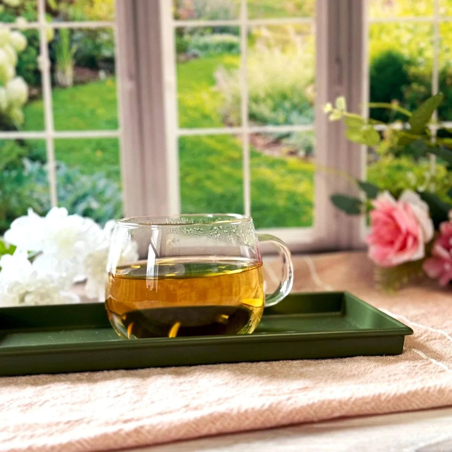 a glass cup of white tea on a tray in front of a window with a spring garden in the background