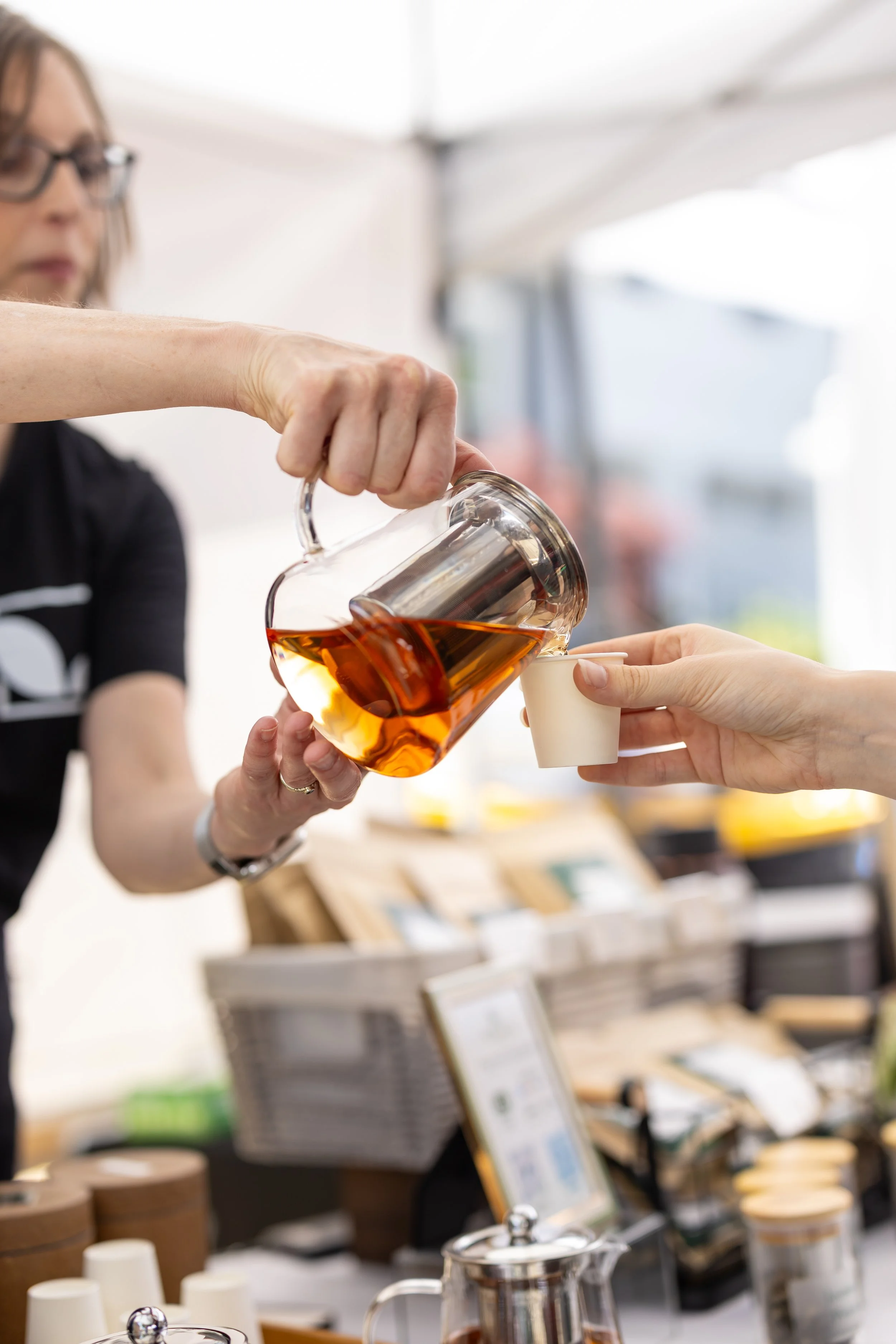 A woman pouring tea from a glass teapot into a small paper cup