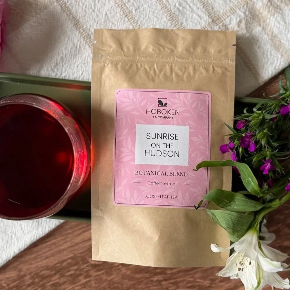 package of Sunrise on the Hudson herbal hibiscus tea from Hoboken Tea Company on a tray next to a cup of hibiscus tea and flowers