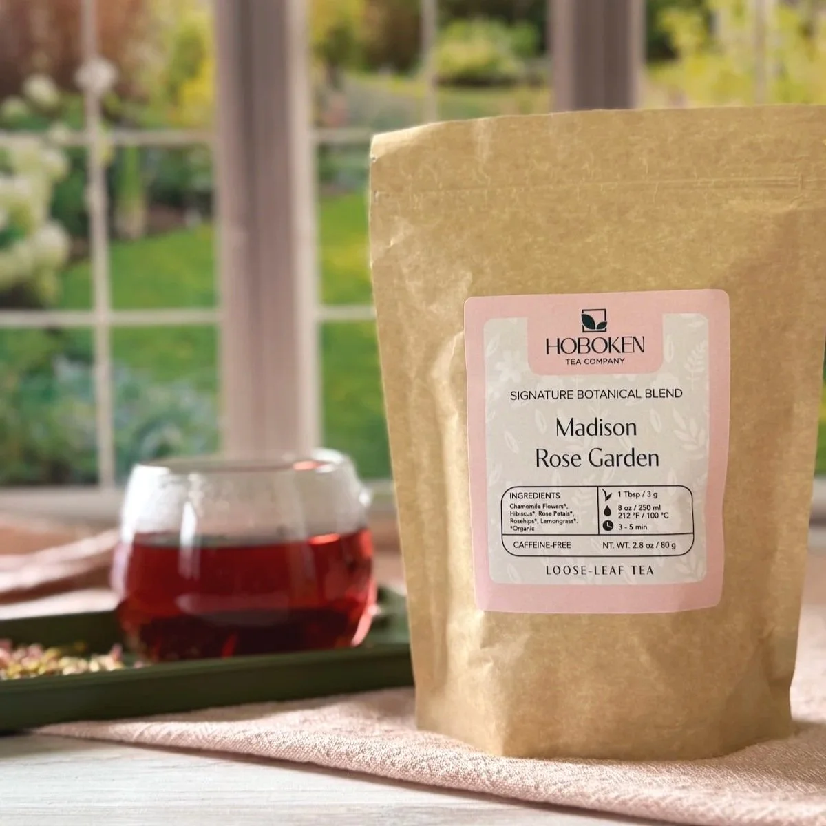 brown paper pouch of Madison Rose Garden herbal tea on a tray next to a cup of pink tea in front of a sunny window with a garden outside