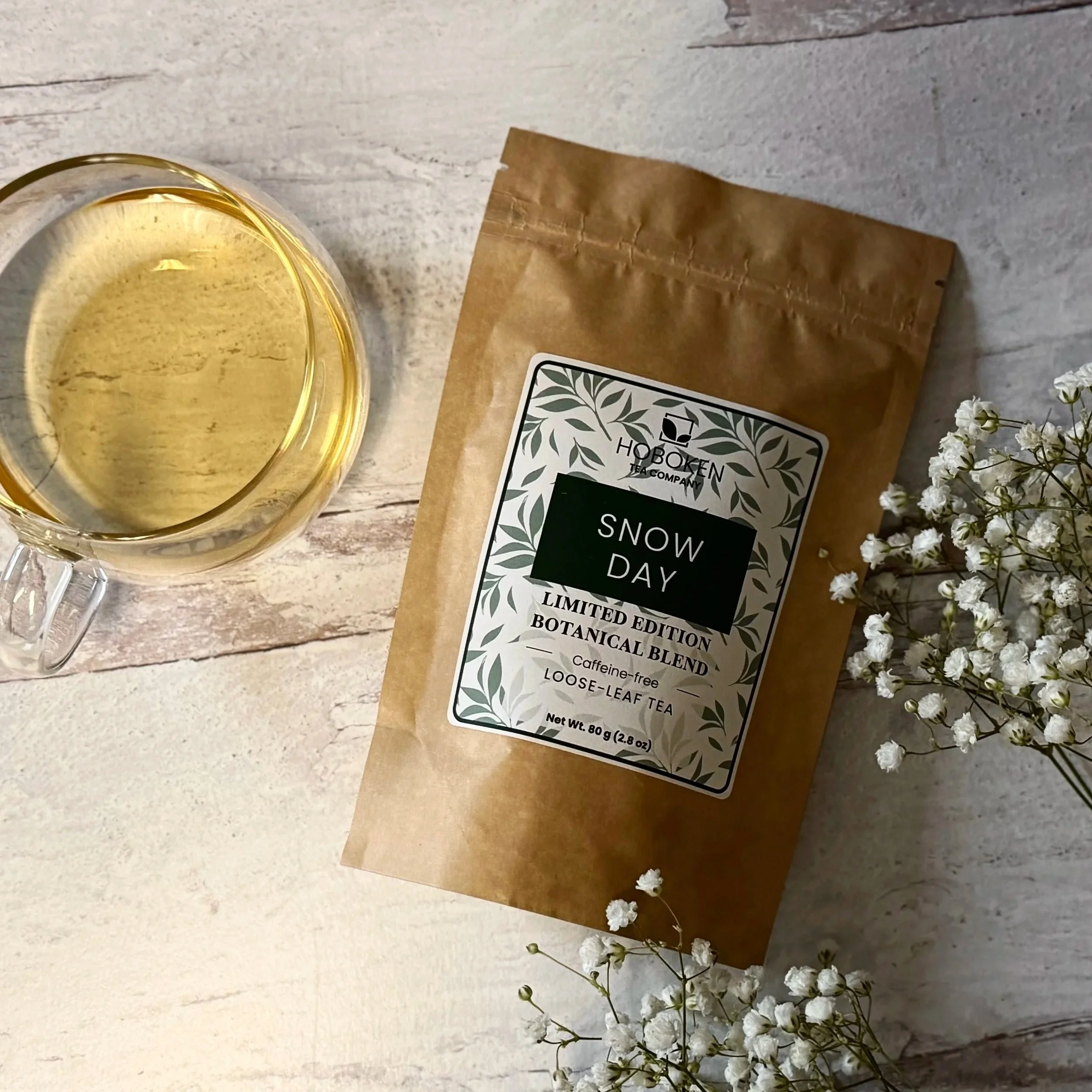 a package of Snow Day loose leaf herbal tea from Hoboken Tea Company next to a glass mug of herbal tea on a light wood table with white flowers
