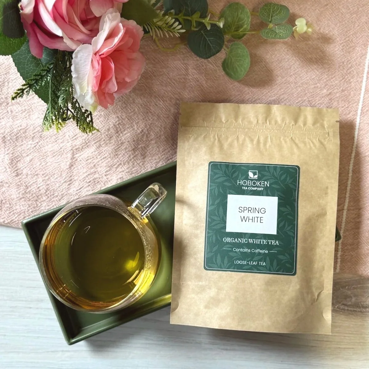 a packet of loose leaf white tea on a tray next to a cup of white tea and some pink flowers