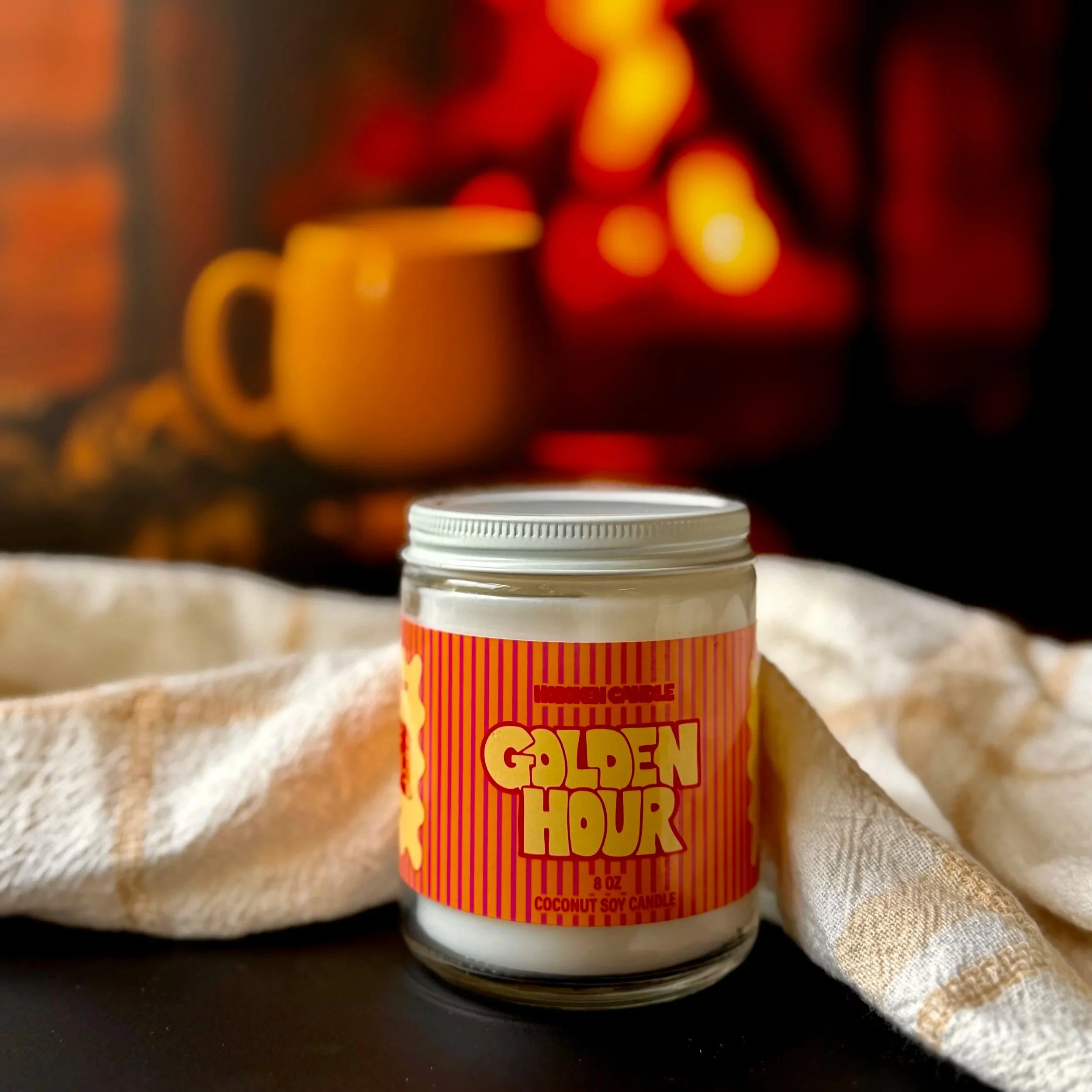 A glass jar labeled Golden Hour with a Hoboken Candle label in red and yellow on a black surface with a fireplace and mug of tea in the background
