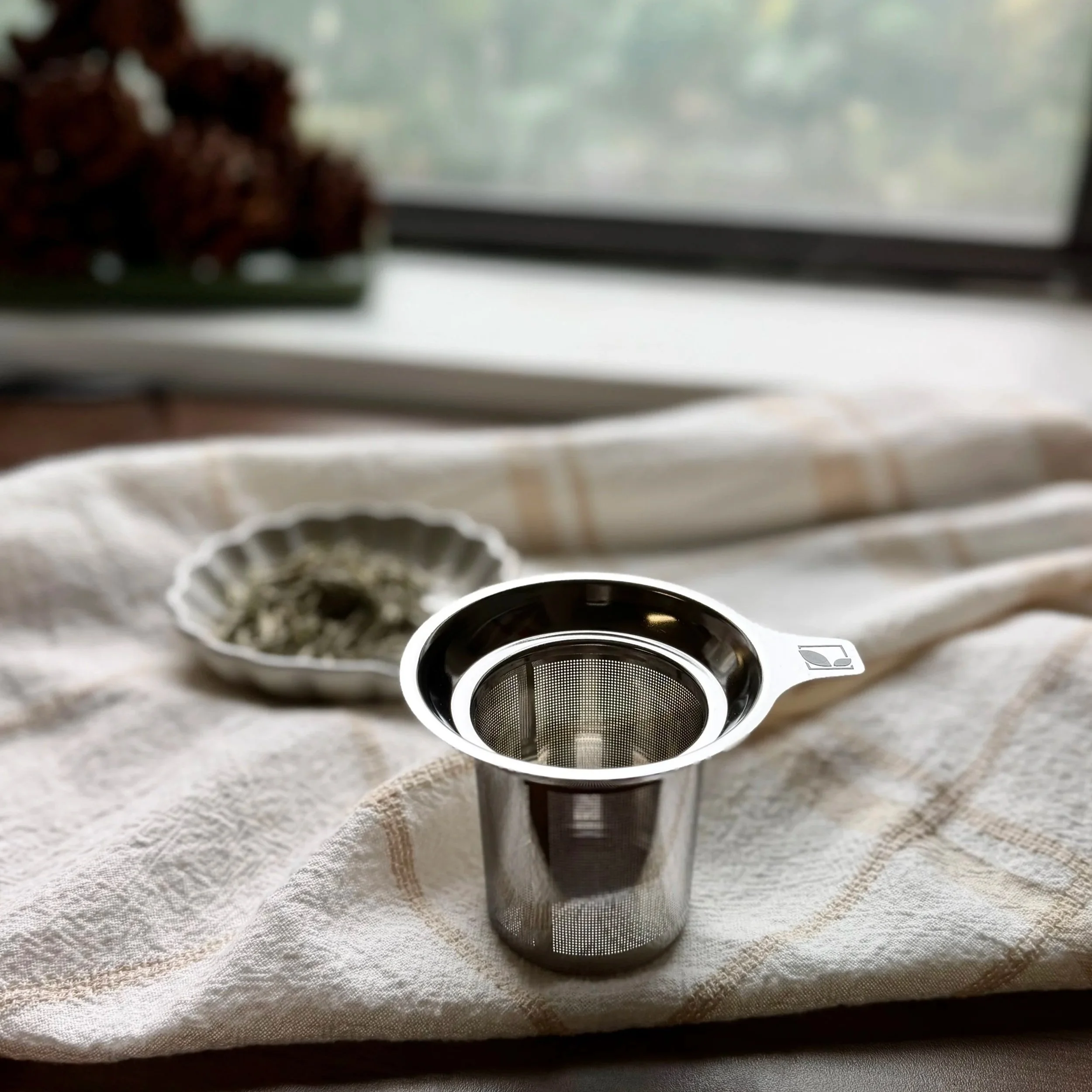 a stainless steel tea infuser basket on a linen towel in front of a window with a bowl of dry tea leaves in the background