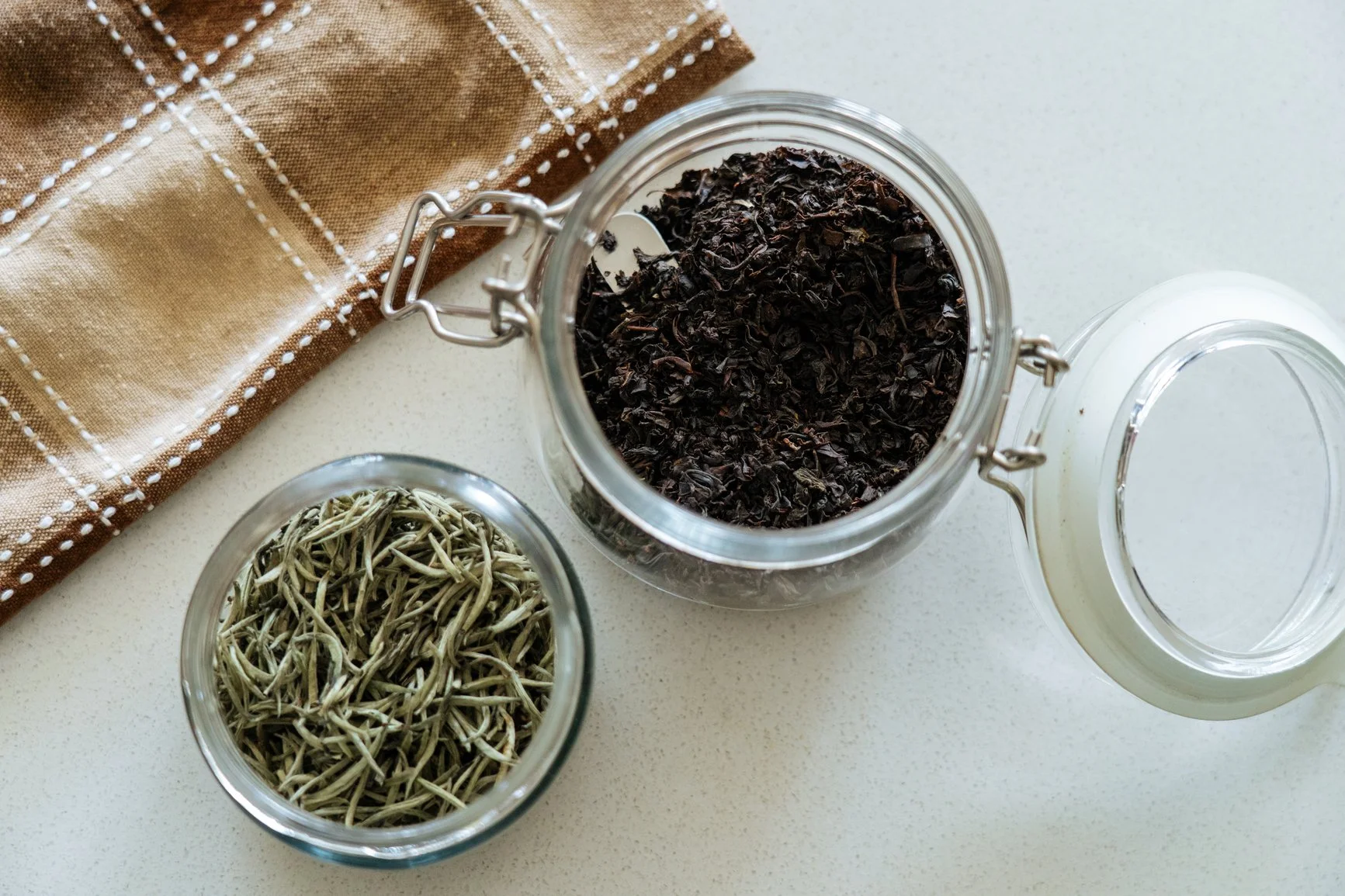 close up of a jar of loose leaf black tea next to a cup of silver needles white tea on a white surface
