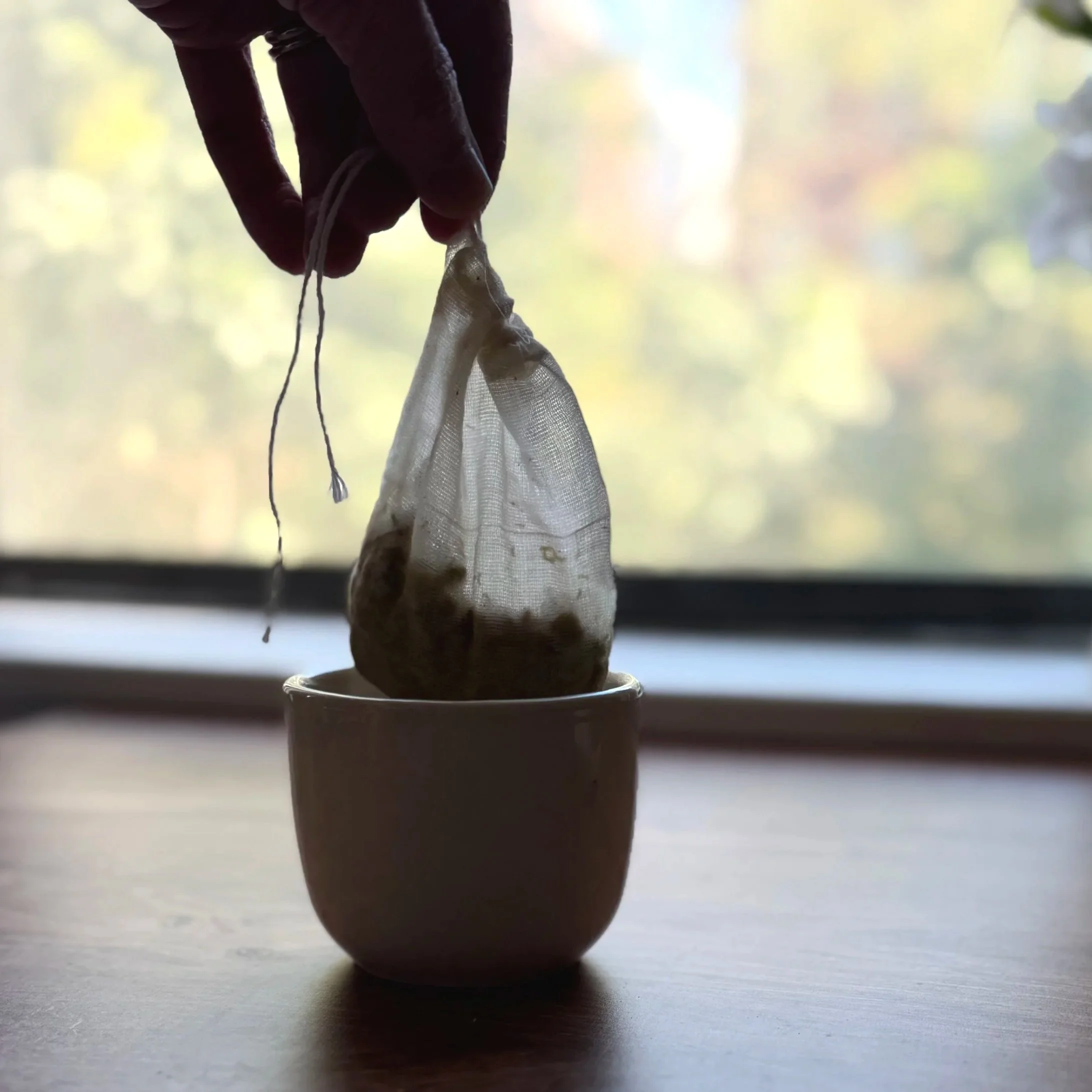 a hand removing a cotton tea bag filled with herbs from a cup of tea