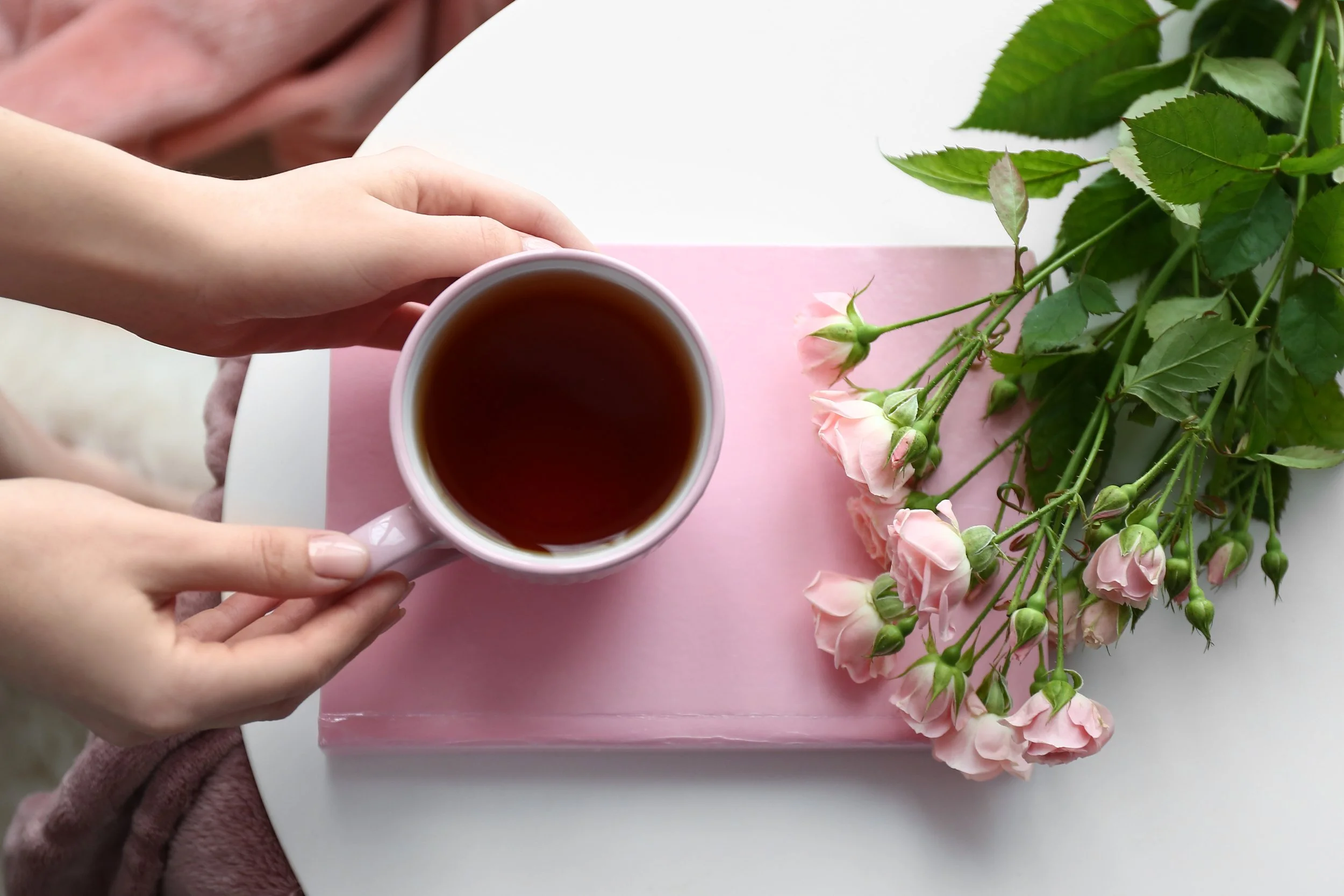 hands holding a cup of tea next to pink flowers and a pink book