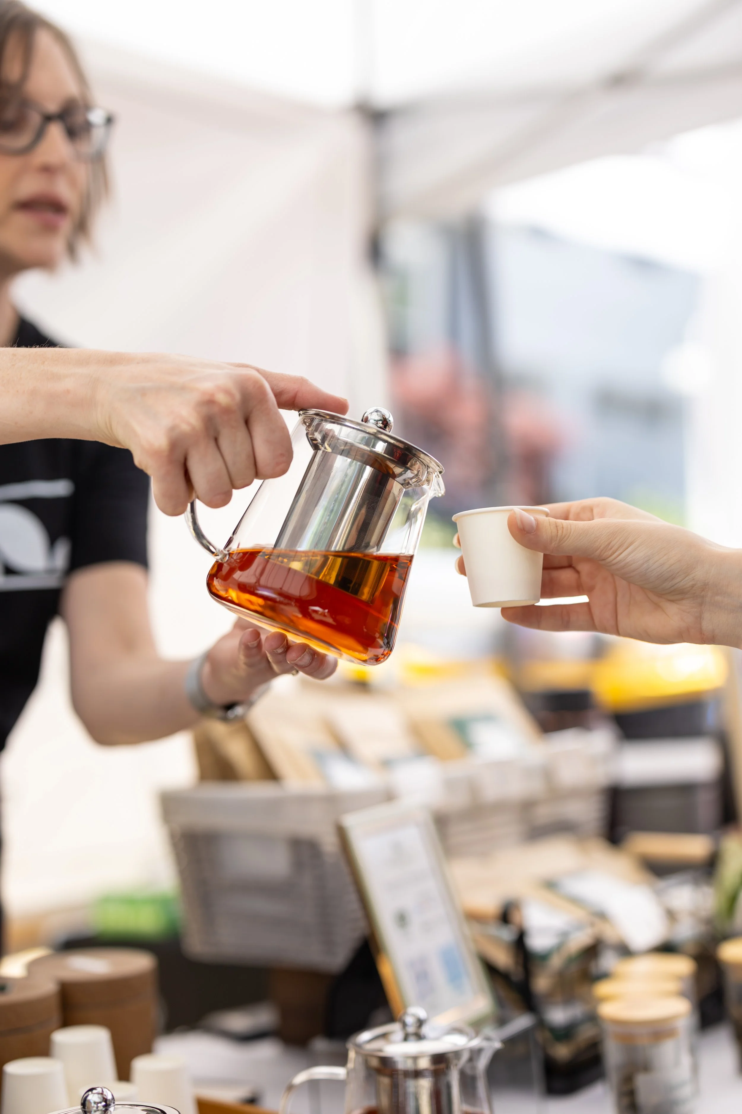 a person pouring tea into a small paper cup from a glass teapot at a market stall