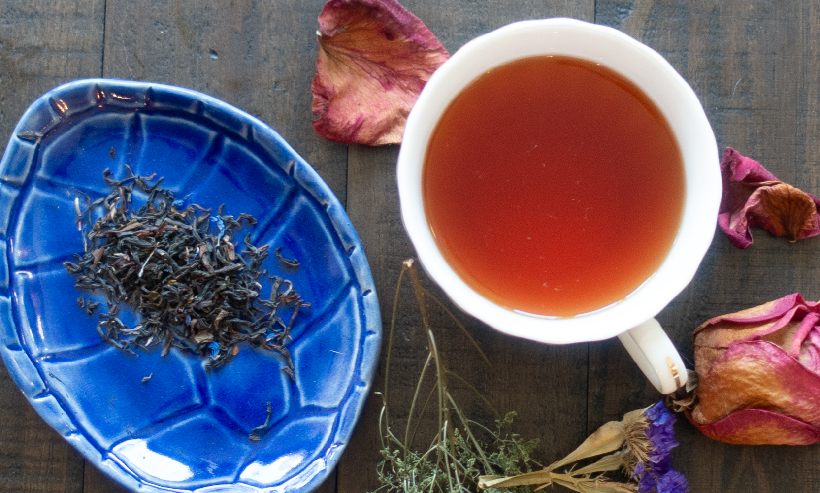 a blue dish with a small pile of loose leaf black tea next to a cup of black tea on a wooden table scattered with dry flowers