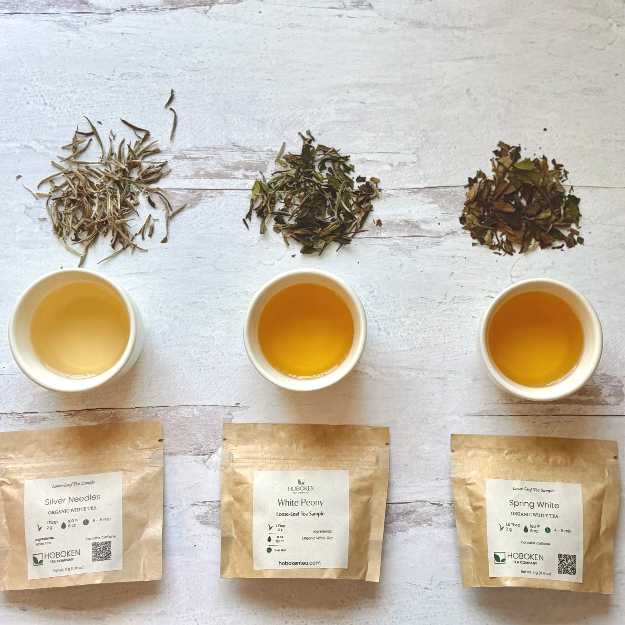 3 small packets of loose leaf white tea with Hoboken Tea Company branding next to cups of tea and piles of dry tea leaves