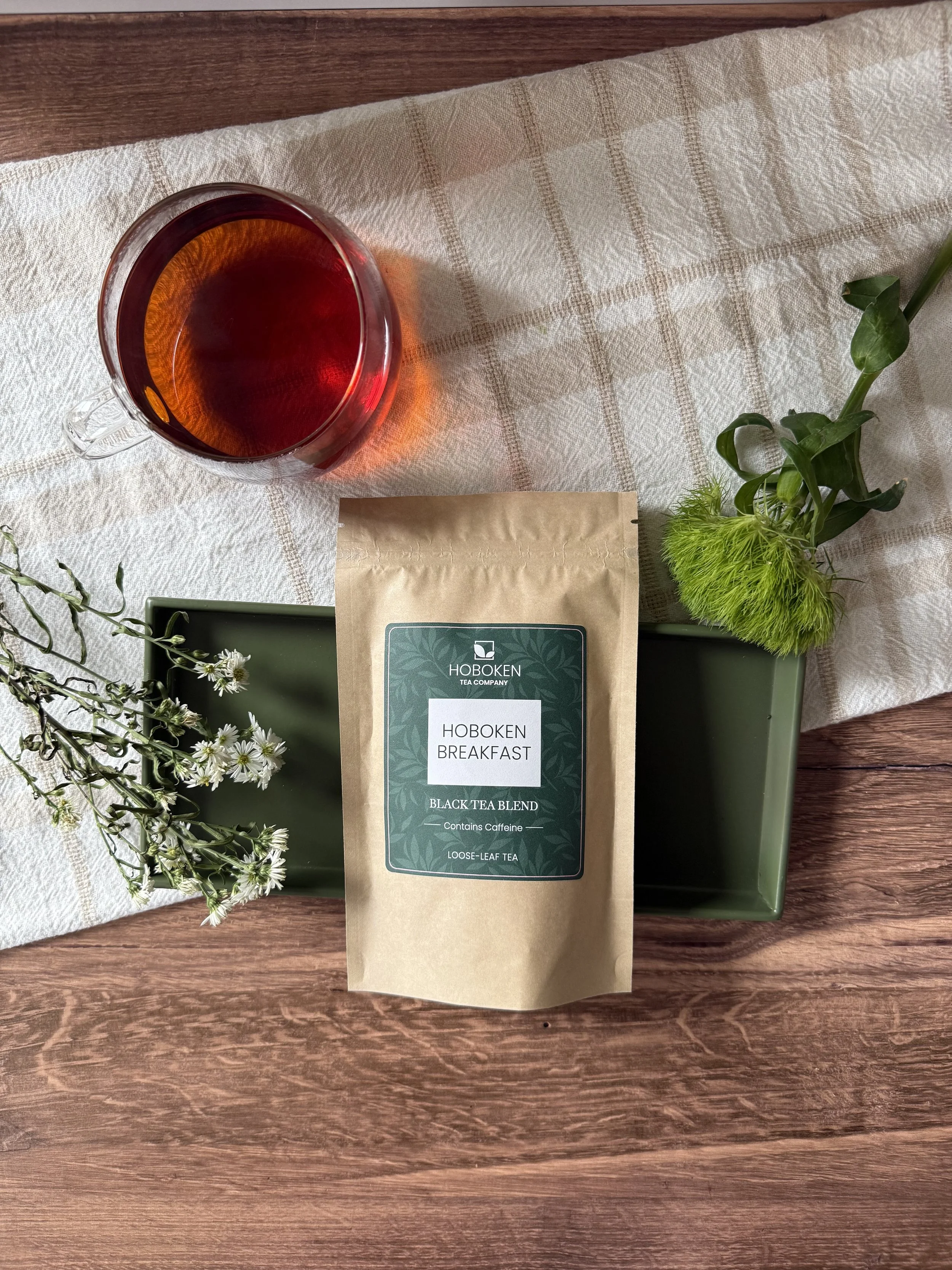 a package of Hoboken Breakfast loose leaf black tea on a wood table next to dried flowers and a glass mug of black tea