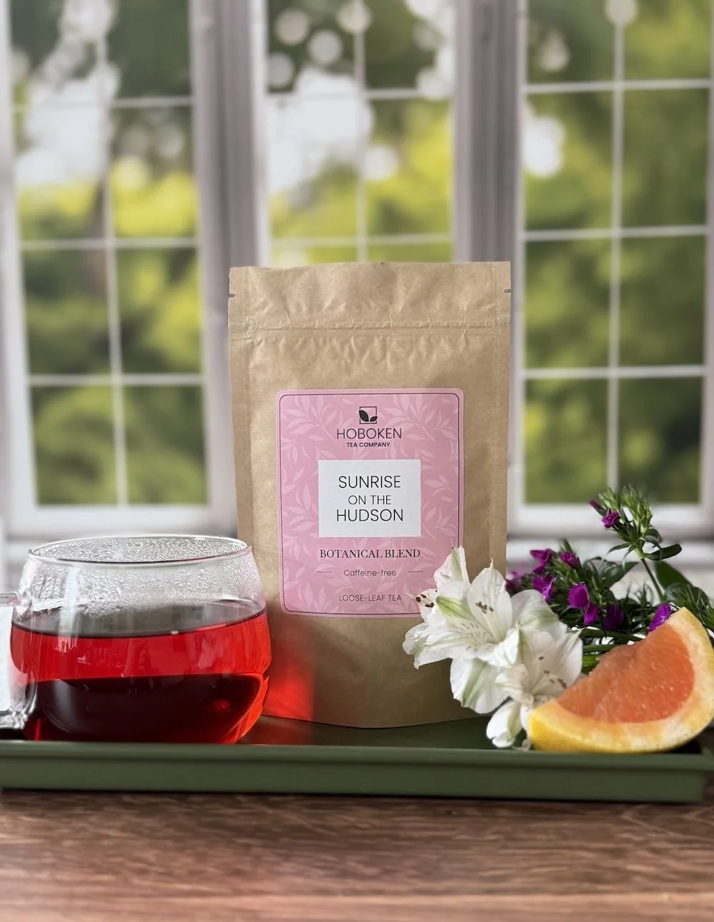Package of Sunrise on the Hudson herbal loose leaf tea from Hoboken Tea Company on a tray with a cup of hibiscus tea, flowers, and fruit in front of a window