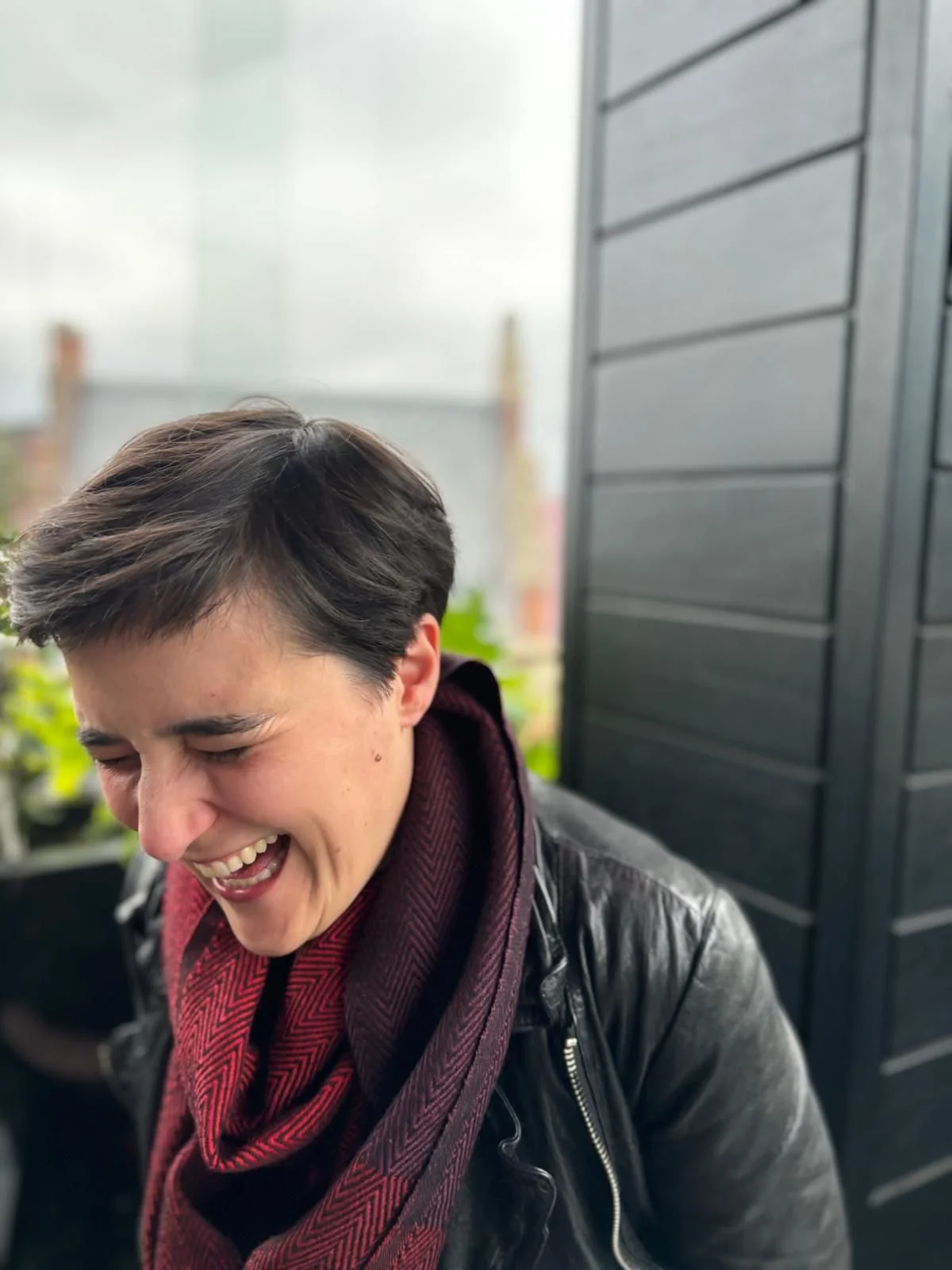 A woman with short dark hair laughing with her eyes closed, wearing a black leather jacket and a maroon scarf, outdoors near a black fence and some blurred greenery.