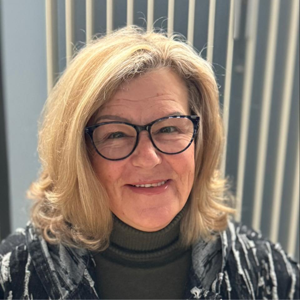 A middle-aged woman with blonde hair, glasses, and a black and white patterned shirt smiling at the camera in front of a gray metal fence.