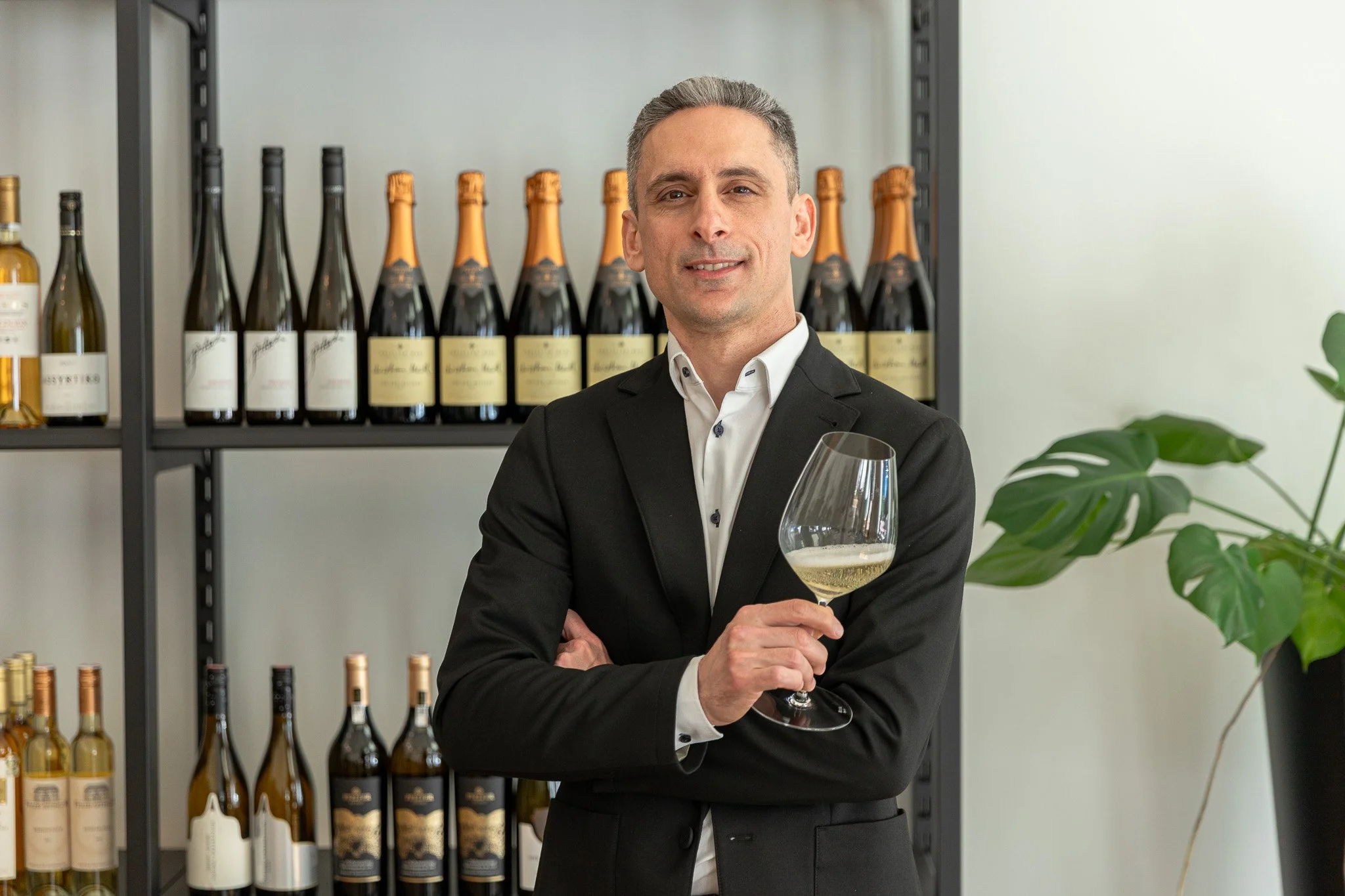 A man in a black suit holding a glass of white wine in a wine cellar.