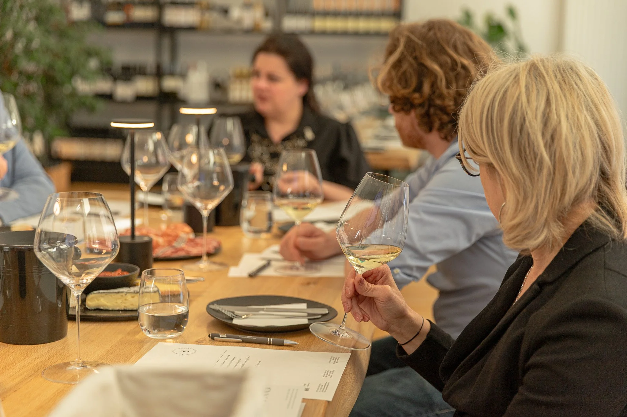 People sitting at a wooden table during a wine tasting or dinner event, holding wine glasses, with some plates of appetizers and paper menus on the table.