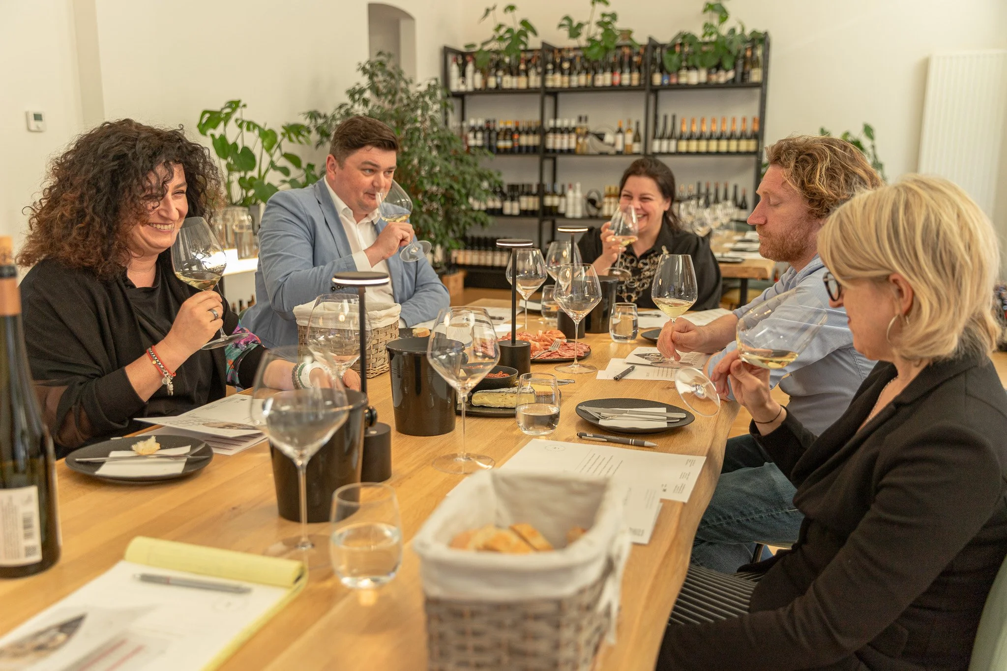 People sitting around a large table enjoying a wine tasting, smiling and smelling wine glasses in a cozy room with a wine rack and greenery in the background.
