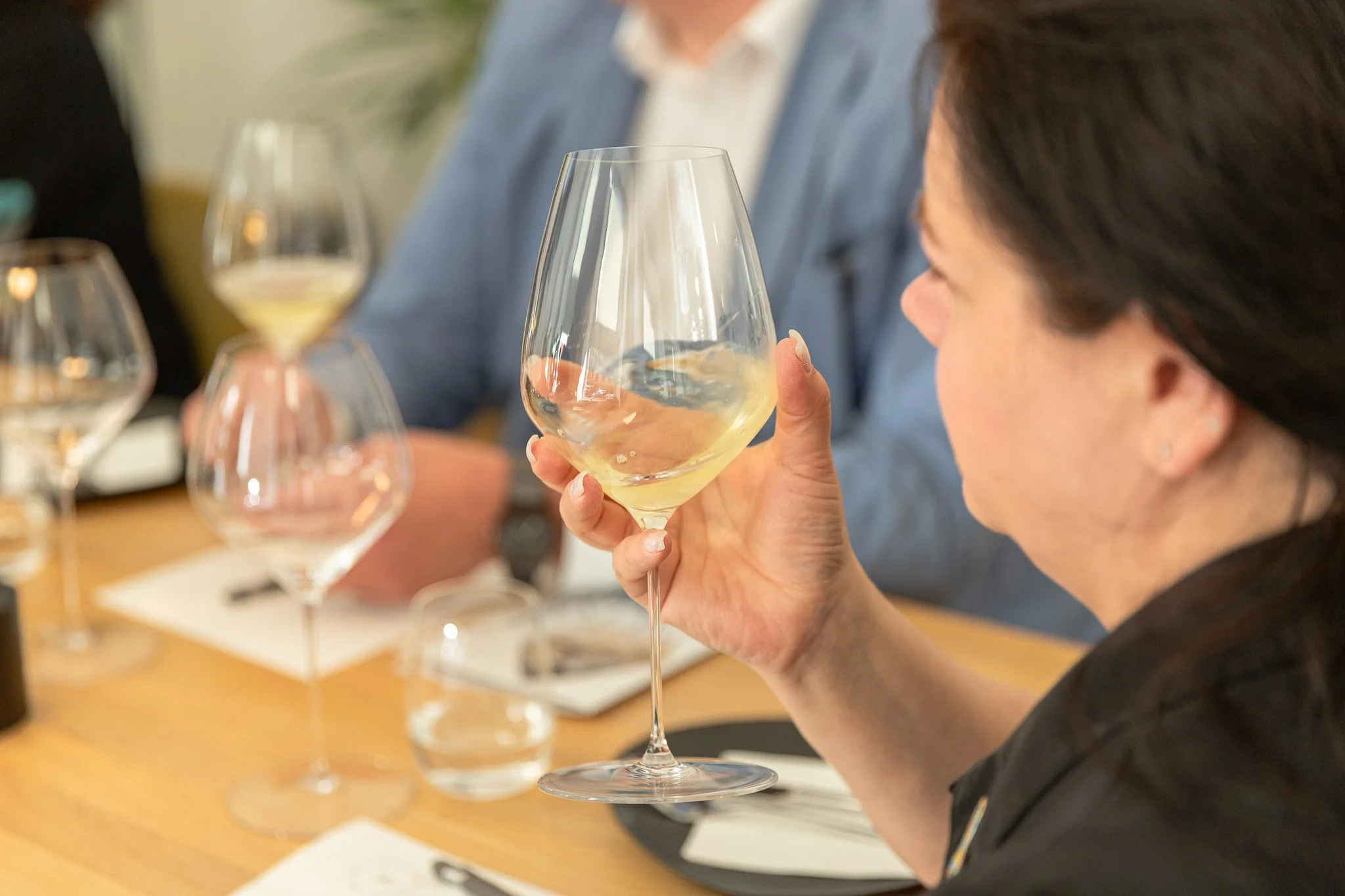 Woman holding a glass of white wine during a tasting, with other glasses and people at the table.
