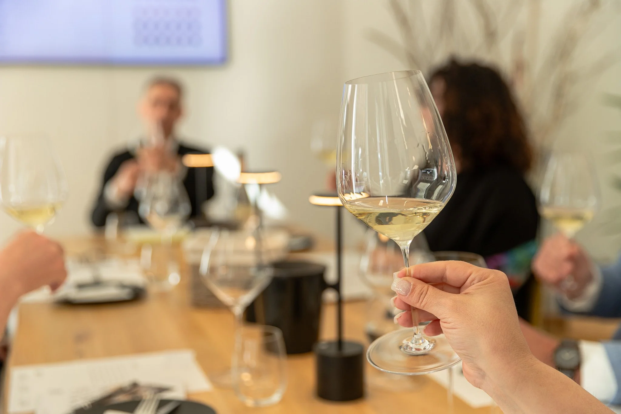 Close-up of a person holding a glass of white wine at a dining table with other wine glasses and people in the background.