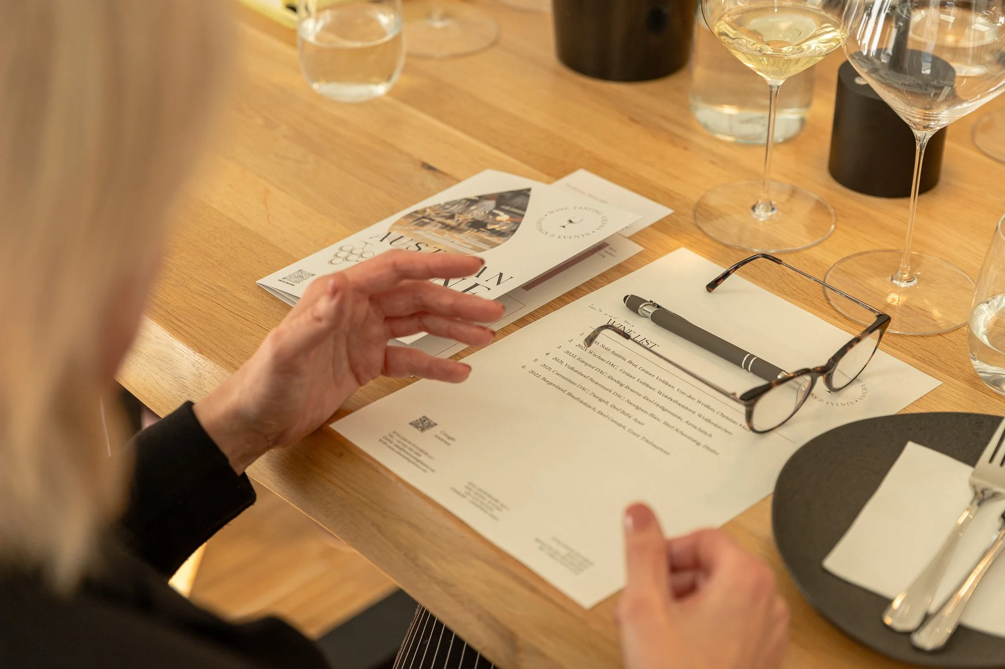 A person preparing to attend a formal event with a wine list, glasses of white wine, cutlery, and a printed menu on a wooden table.