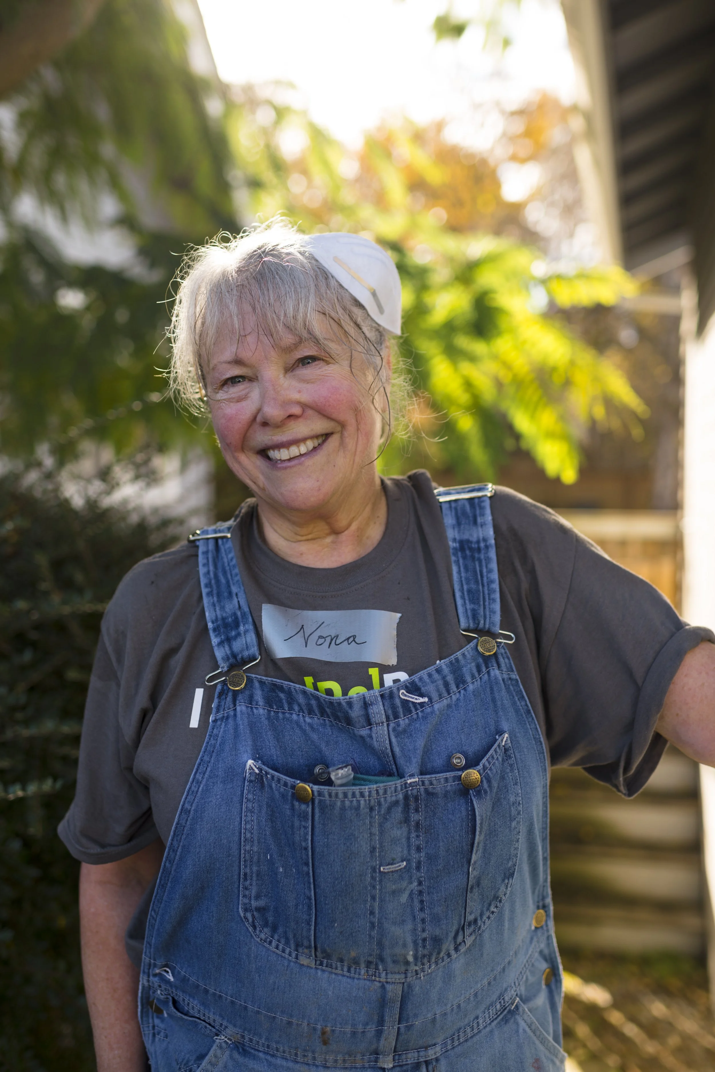A smiling woman with gray hair and a name tag that says 'Nona' standing outdoors in sunlight, wearing a gray t-shirt and blue denim overalls, with a green leafy background.