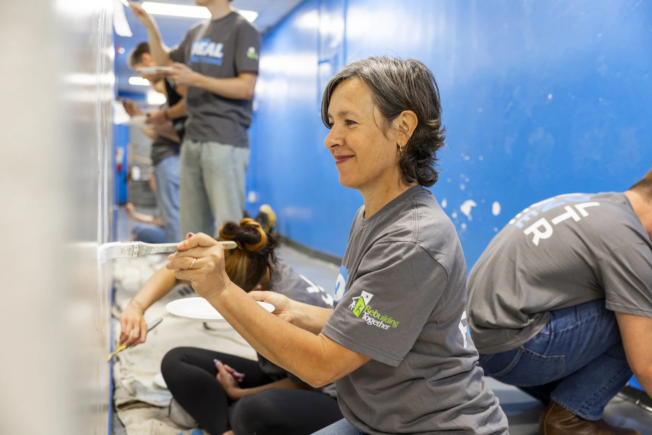 Woman sitting on the floor, smiling, holding a white plate and a fork, participating in a community event, with others in the background near blue walls.