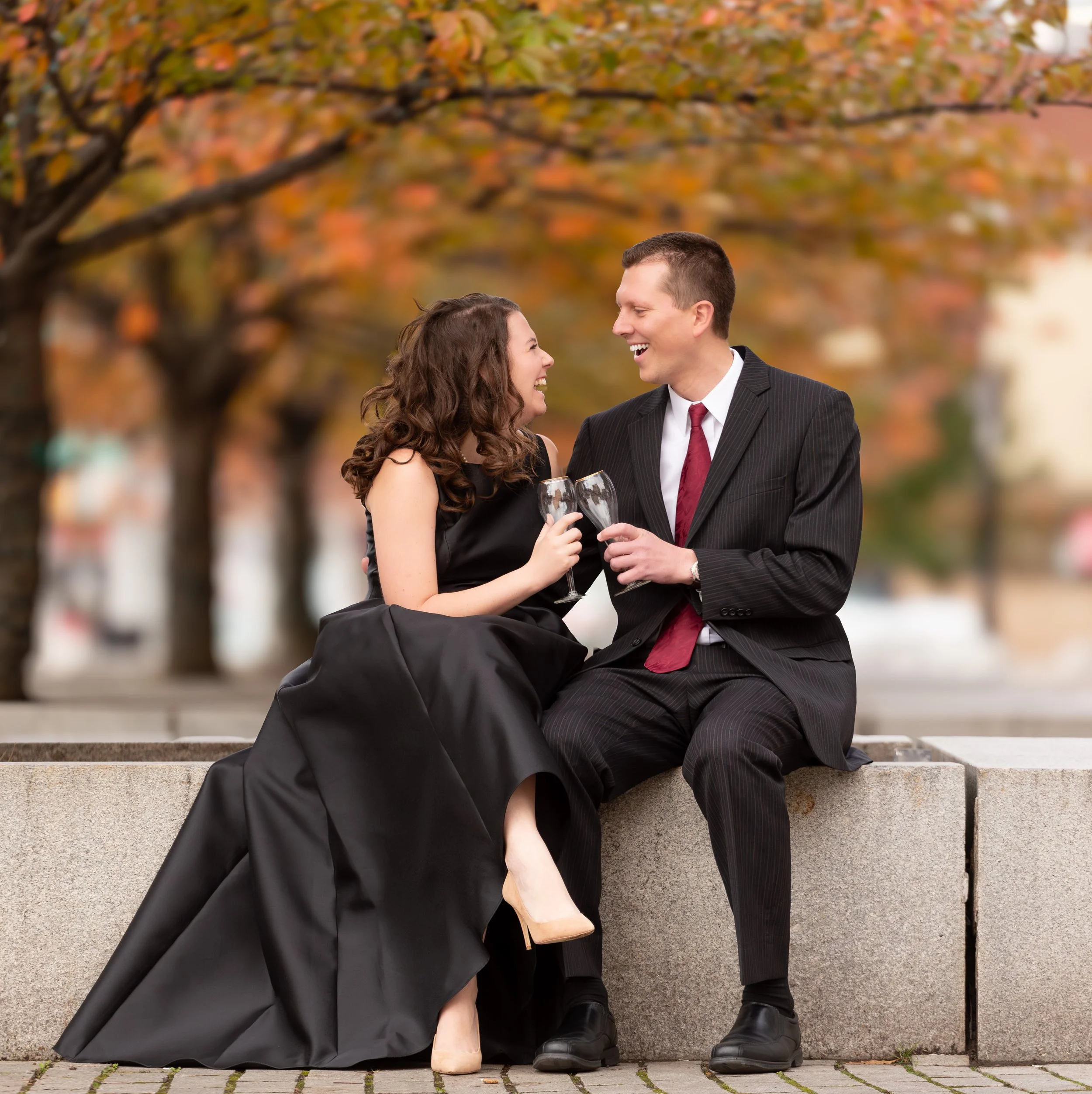 A couple dressed in formal attire sitting on a concrete bench outdoors, holding champagne glasses, smiling at each other against a backdrop of autumn trees.