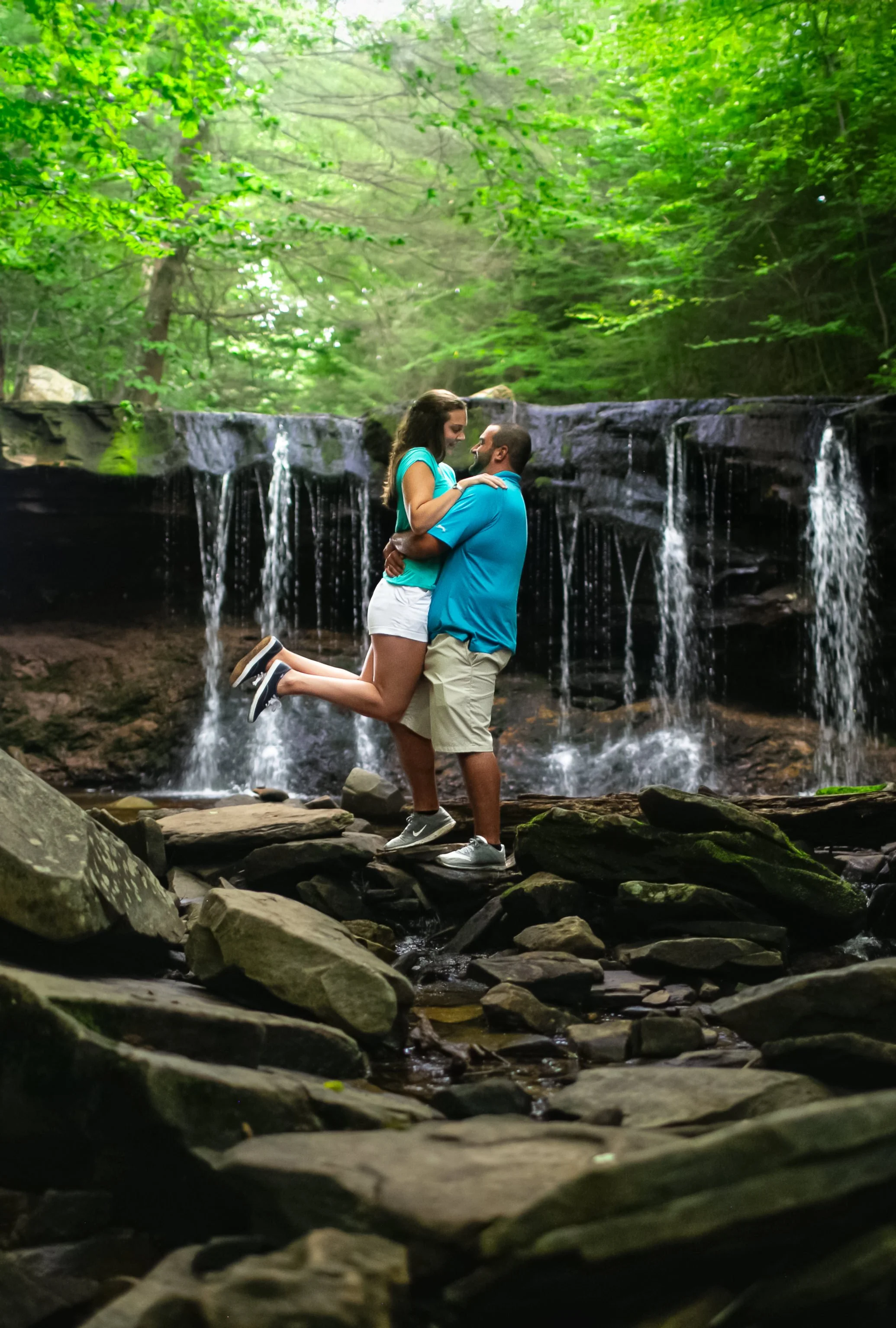 A couple embracing near a waterfall in a lush, green forest, with the woman being lifted by the man. The scene is set on rocks at the edge of the stream.